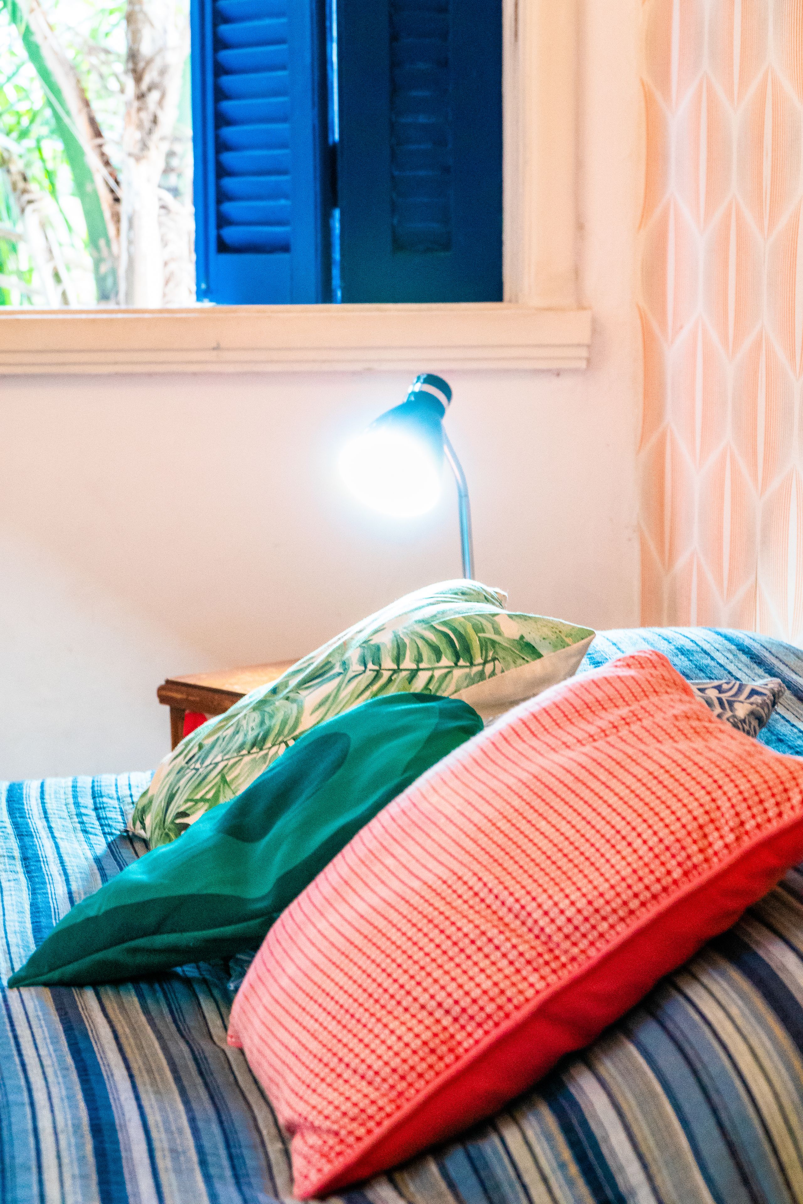 A bed with striped bedding and two pillows, a bedside lamp, and a window with blue shutters.