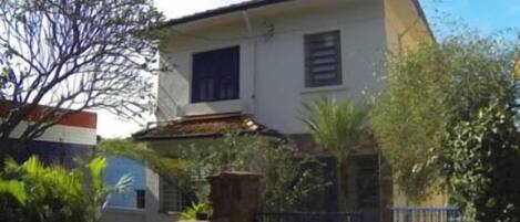 Una casa de dos pisos con fachada blanca, un ventanal en el nivel superior y un muro de ladrillo al frente.