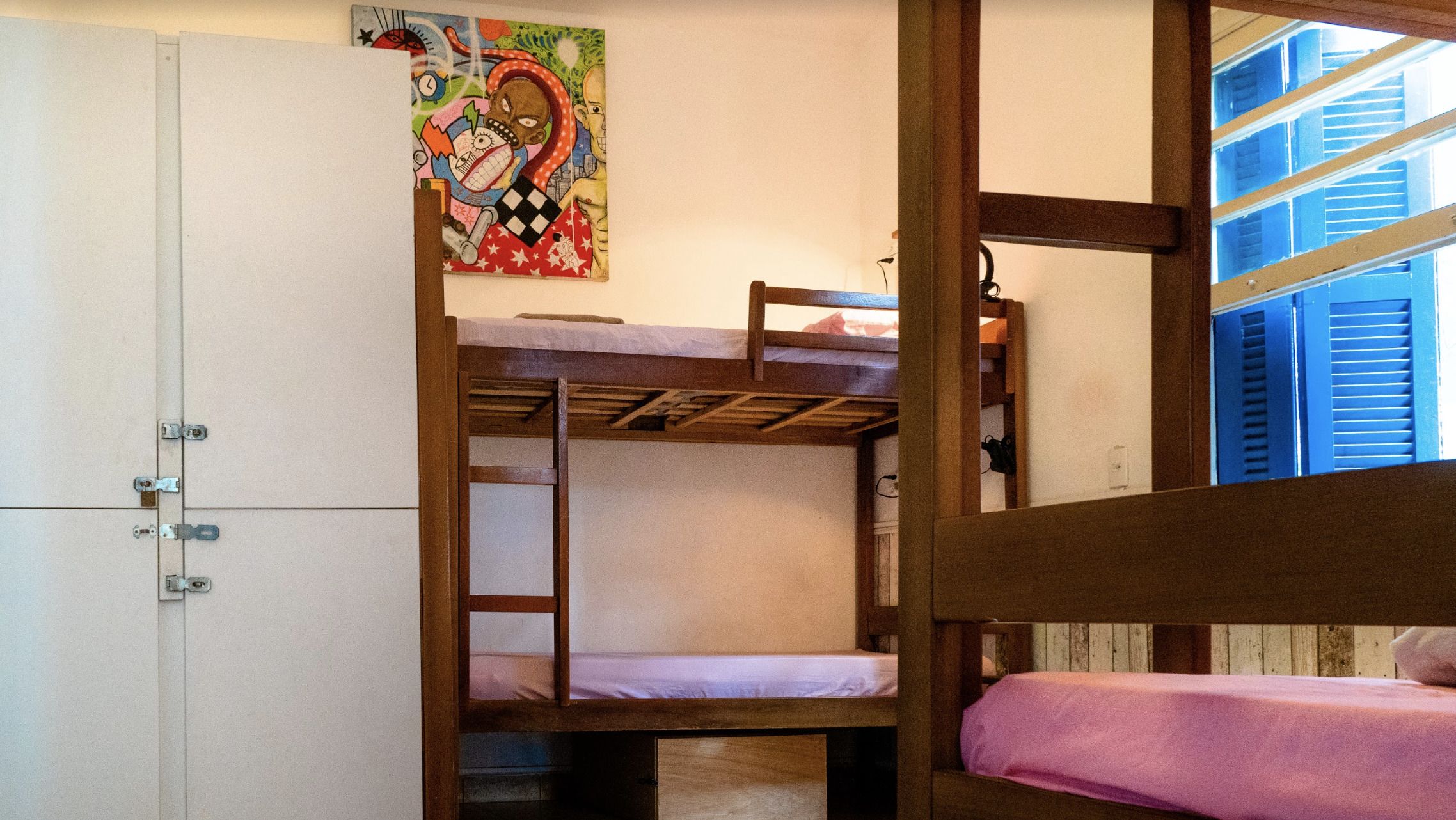 Habitación con literas de madera, un armario blanco y un cuadro de colores en la pared.