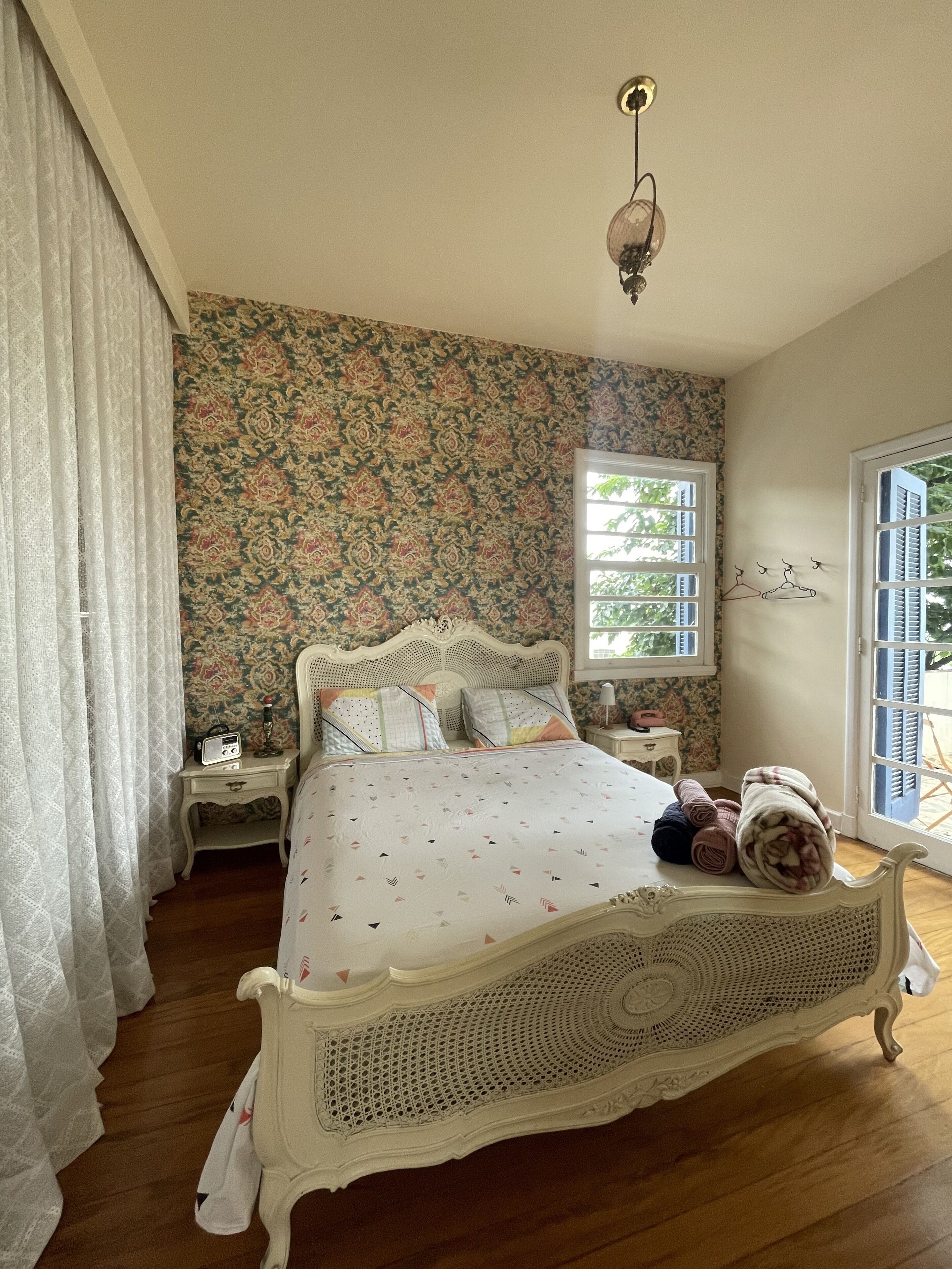 A bedroom with a large bed, floral wallpaper, a window with shutters, and a hanging light fixture.