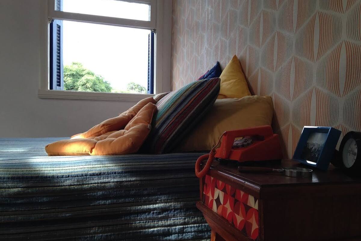 A bed with striped bedding, a window with a view of greenery, and a bedside table with a clock and a picture frame.