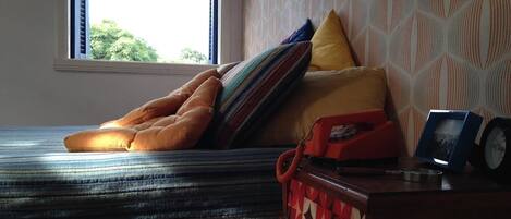 A bed with striped bedding, a window with a view of greenery, and a bedside table with a clock and a picture frame.