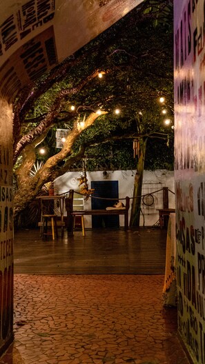An outdoor seating area with tables and chairs under a tree, surrounded by walls with posters. - Paradiso Hostel (São Paulo)