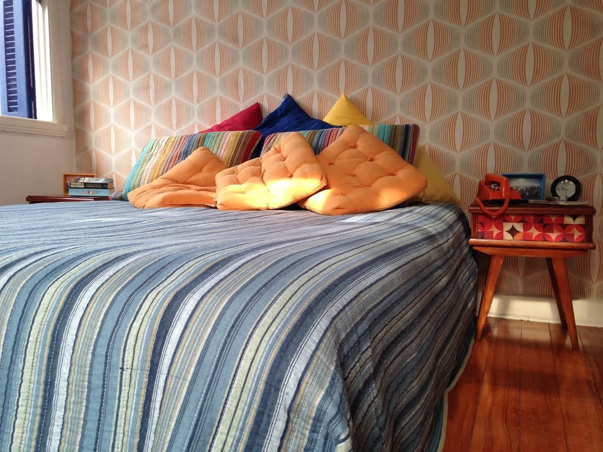 Un dormitorio con colcha a rayas, varios almohadones, una mesita de noche de madera con un reloj y papel tapiz estampado.