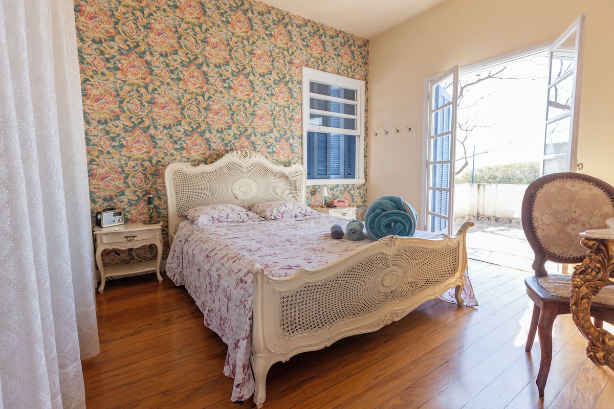 A bedroom with a floral wallpaper, a white ornate bed, a wooden floor, and a view through an open window.