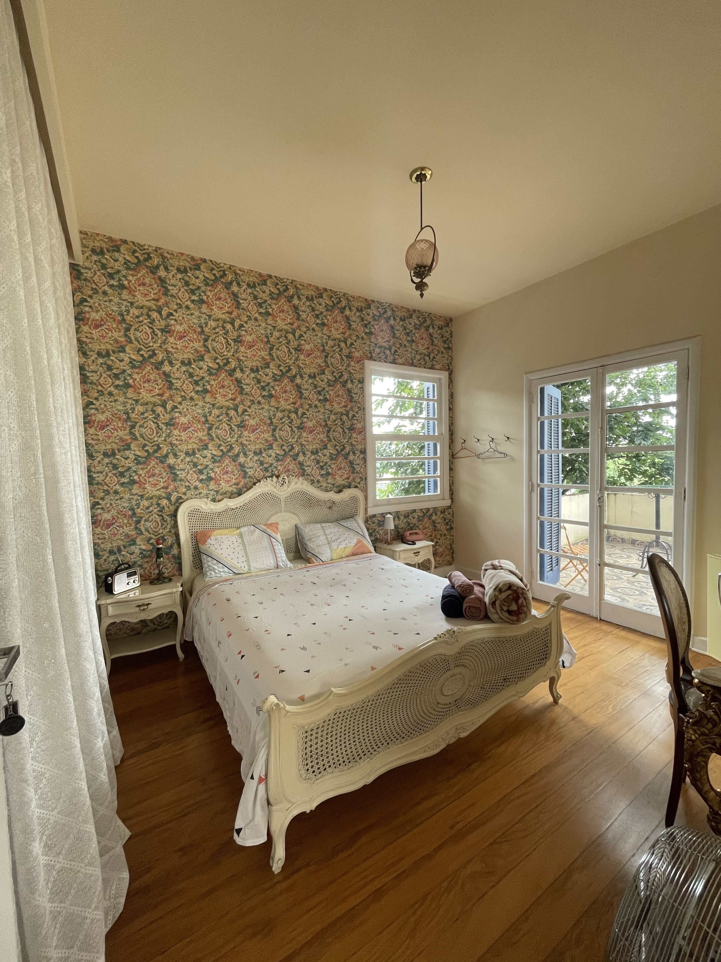Un dormitorio con papel tapiz floral, una cama clásica, una mesita de noche con un radio y vistas a través de un ventanal.