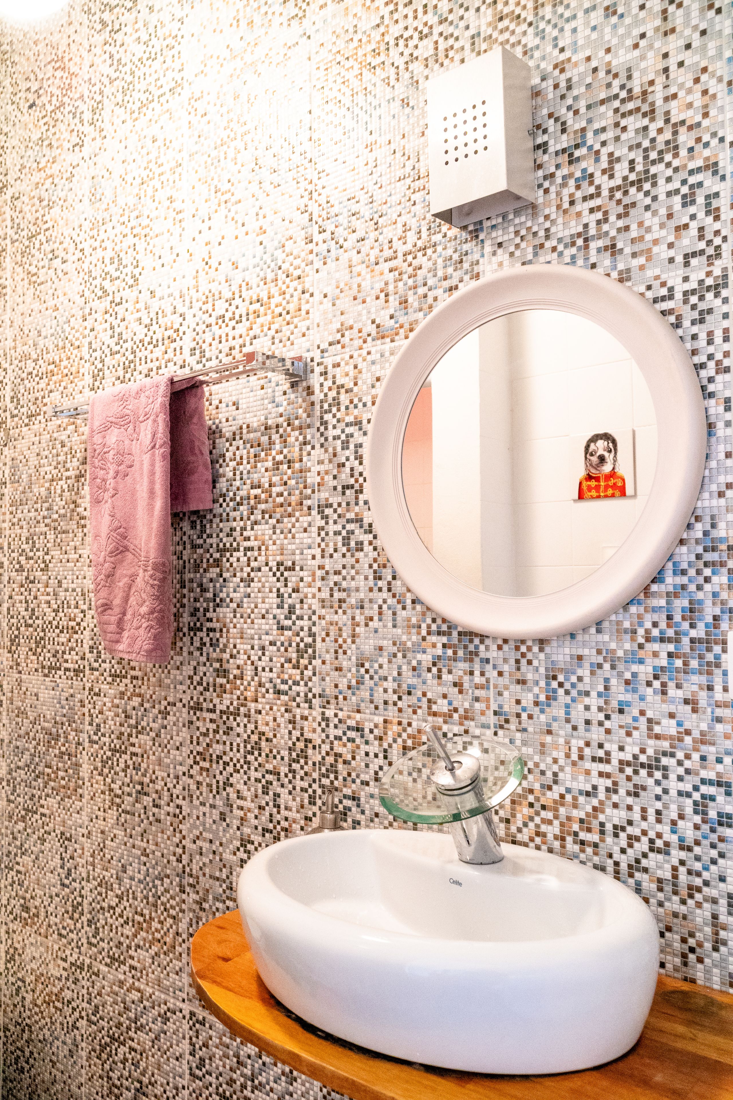 Un baño con pared de mosaicos, un espejo redondo, un lavabo con cubierta de vidrio y una toalla rosa colgada en un perchero.