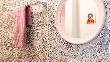Un baño con pared de mosaicos, un espejo redondo, un lavabo con cubierta de vidrio y una toalla rosa colgada en un perchero.