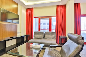 Une chambre d’hôtel moderne avec un canapé, une table basse en verre, un téléviseur fixé au mur et des rideaux rouges.