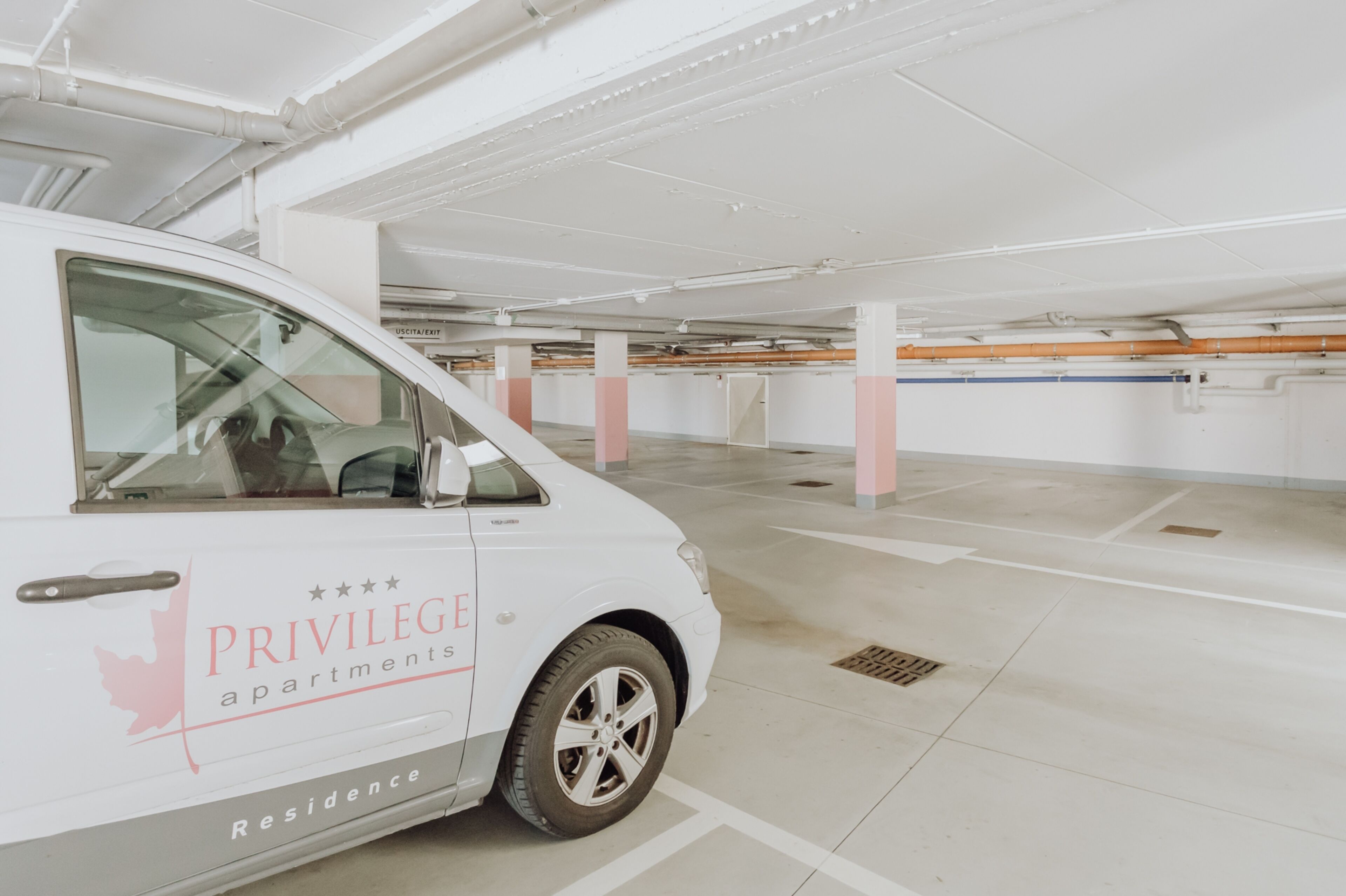Ein weißer Van mit Schriftzug „PRIVILEGE apartments“ steht in einer sauberen, leeren Parkgarage mit weißen Säulen und gefliestem Boden.