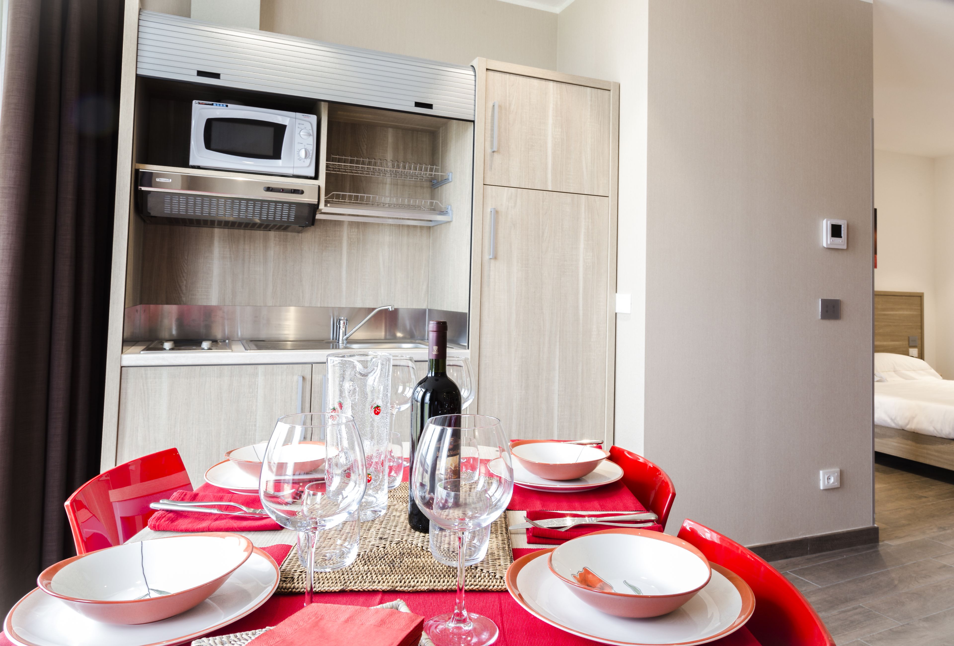 Una habitación de hotel compacta con kitchenette, un comedor con sillas rojas y una mesa dispuesta para dos, y una cama en la habitación contigua.