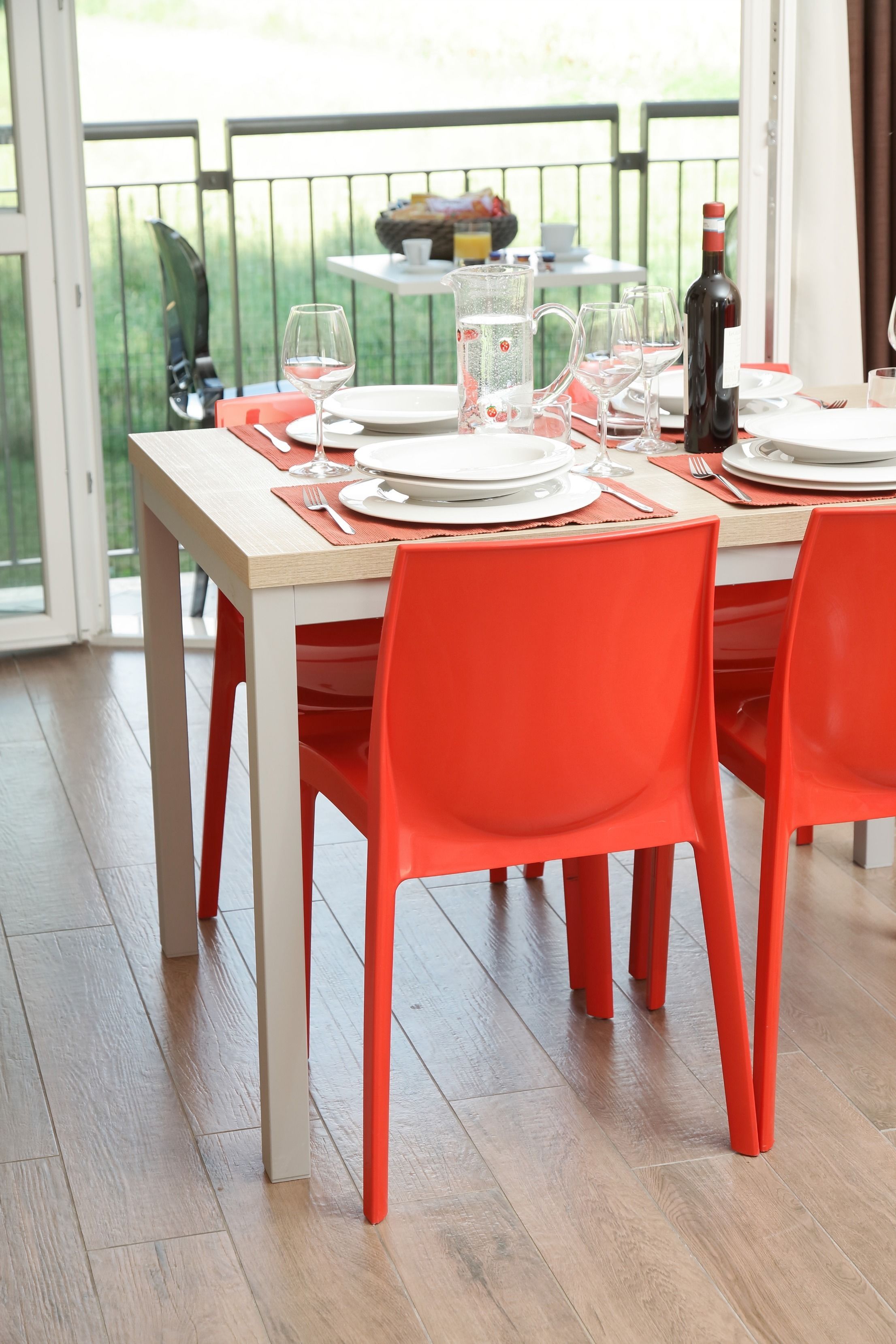 A dining table set with plates, glasses, and cutlery, accompanied by bright orange chairs, in a room with a view of greenery outside.