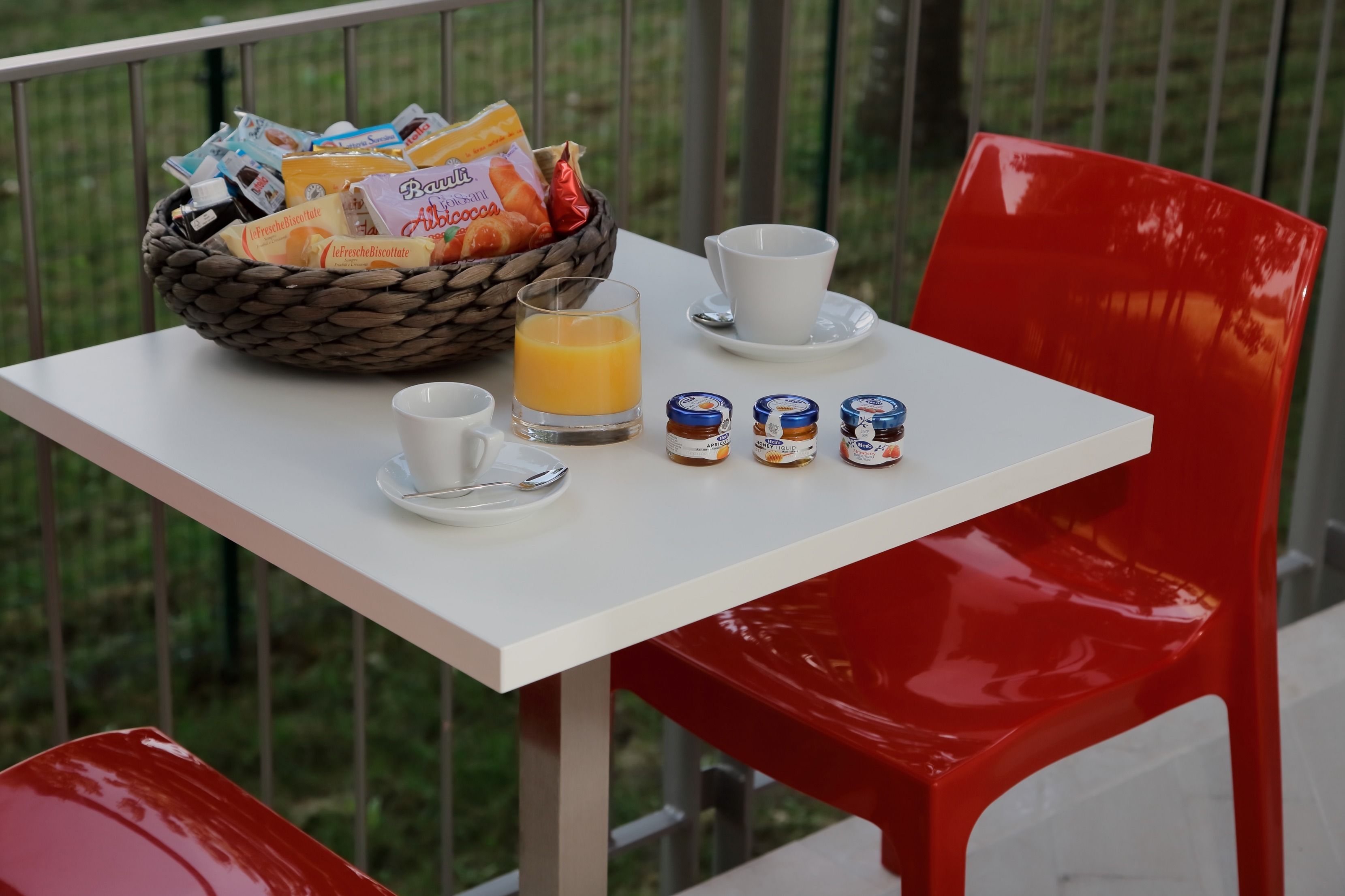 Un desayuno con una cesta de pasteles, un vaso de zumo de naranja y tres botes de mermelada sobre una mesa blanca.