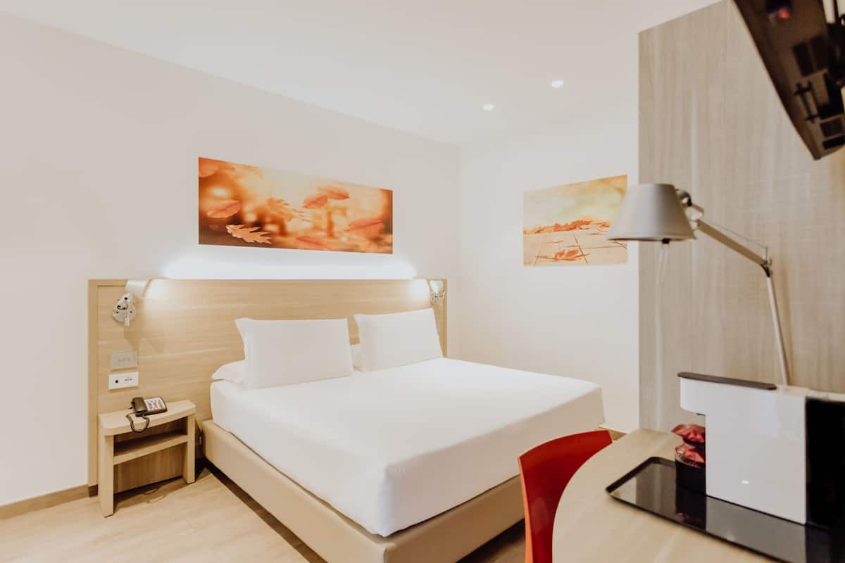 Una habitación de hotel con cama, mesita de noche, lámpara y una obra de arte en la pared.