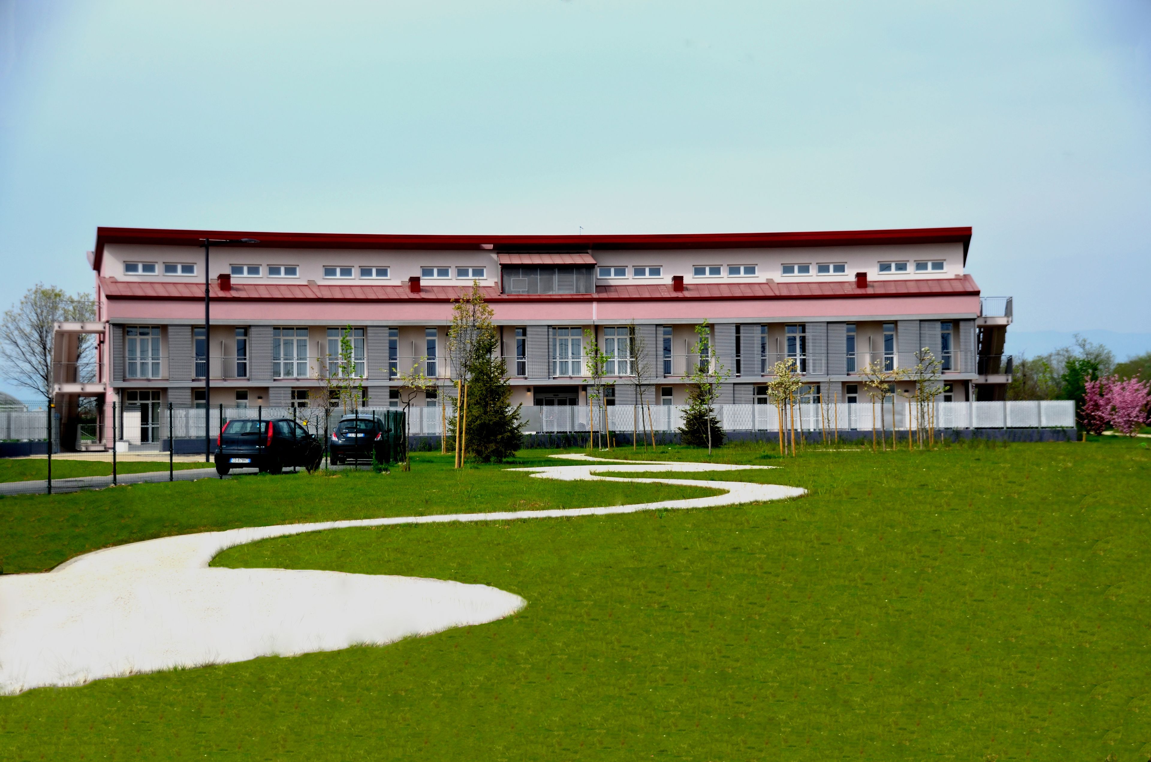 Un edificio grande con tetto rosso e numerose finestre. C'è un prato verde con un vialetto sinuoso e alcuni alberi.