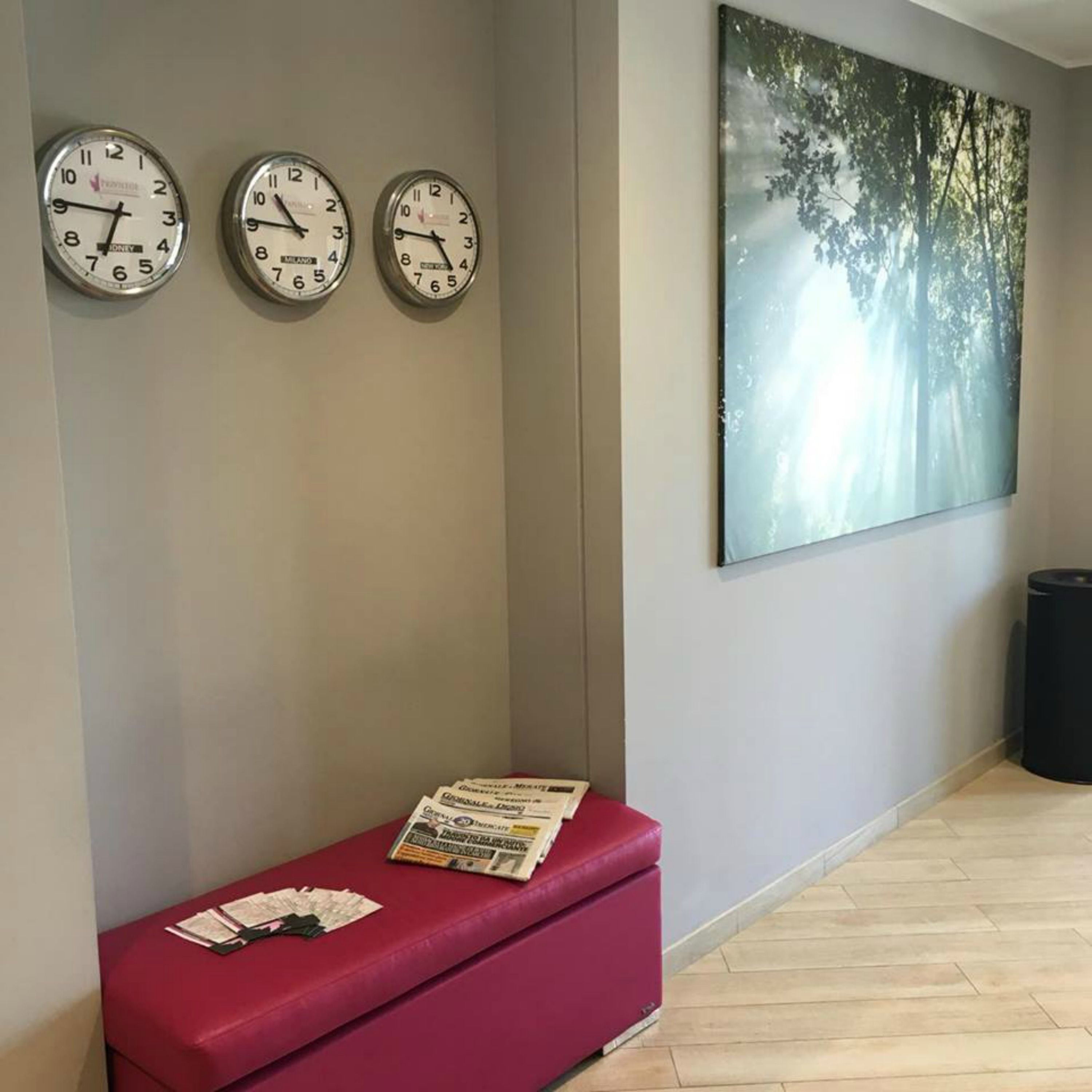 A reception area with three clocks on the wall, a bench with a newspaper, and a large forest-themed painting.