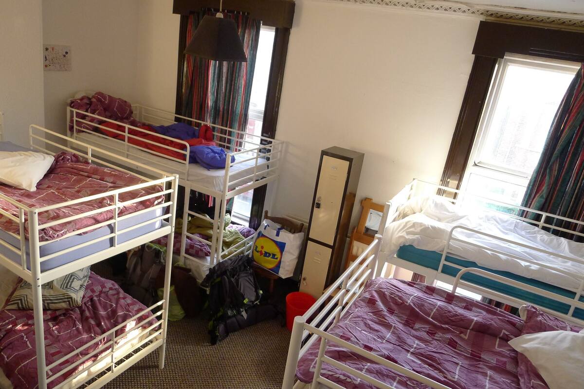 A dormitory room with bunk beds, a wardrobe, a mirror, and a window with curtains.