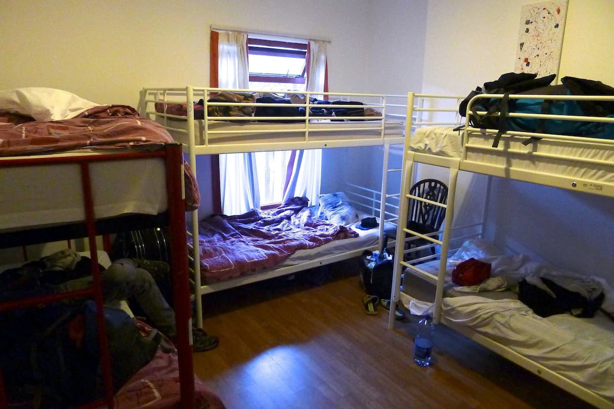 A dormitory room with bunk beds, a window with curtains, and a wooden floor.