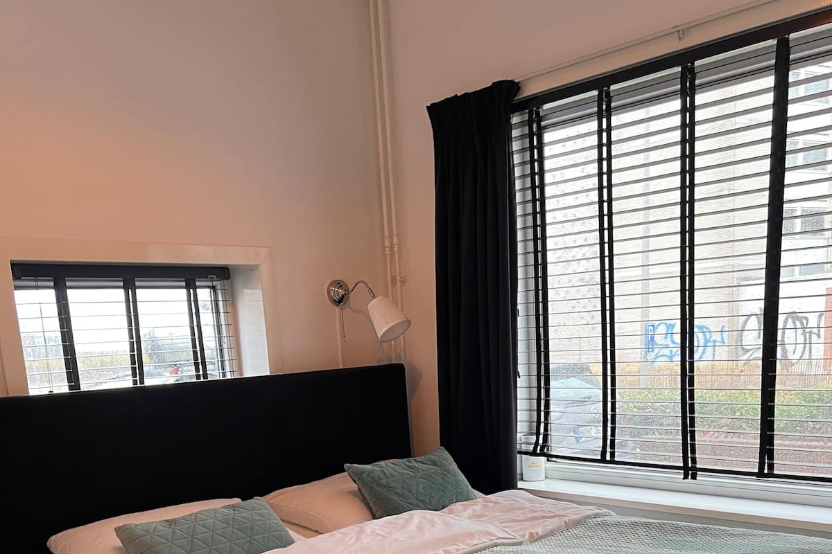 Een net opgemaakt bed met een gewatteerd sprei en kussens, een zwart hoofdbord, en een raam met jaloezieën en gordijnen.