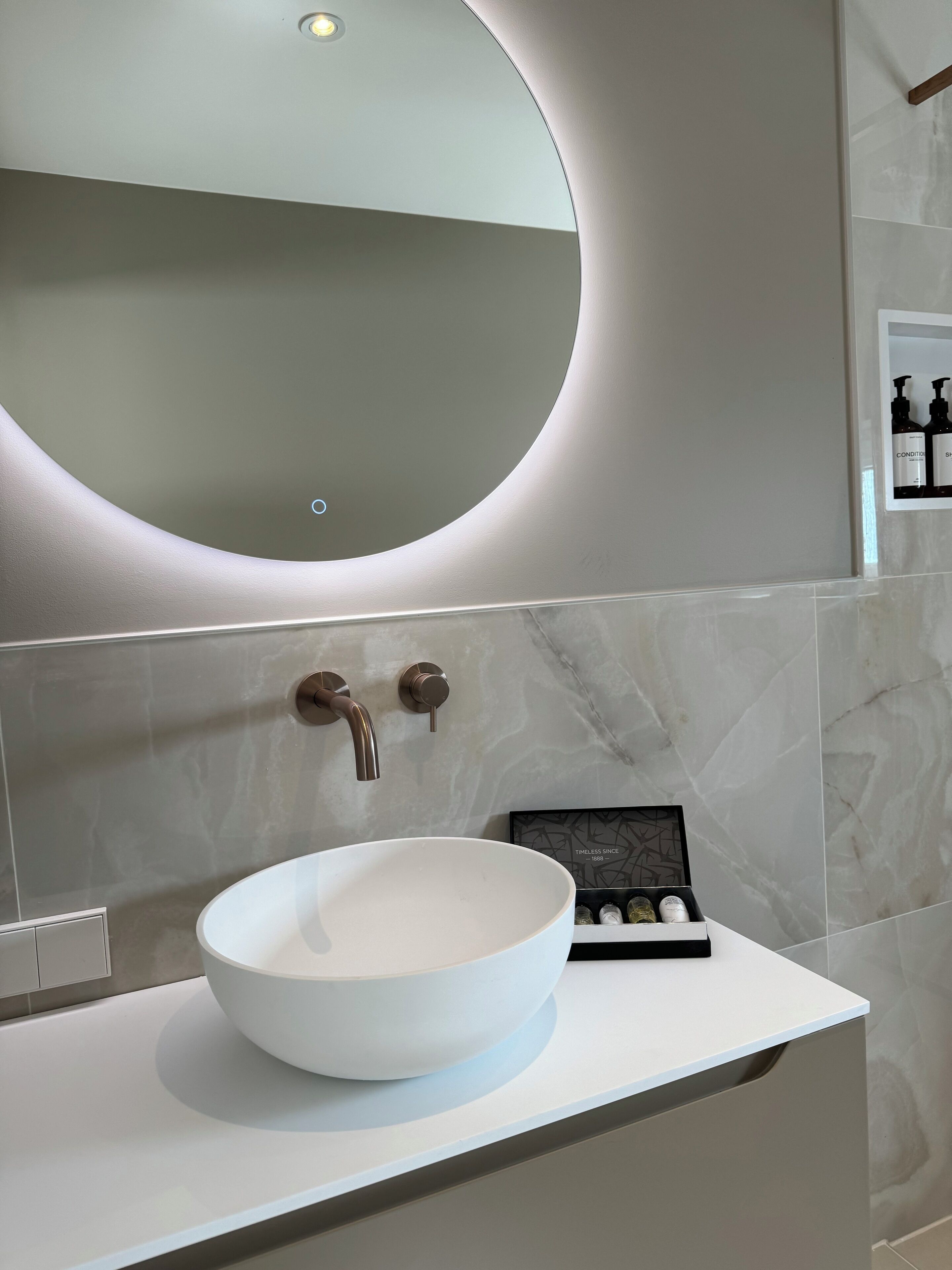 Une salle de bain moderne avec un grand miroir rond, un lavabo sur colonne blanc et un petit présentoir pour les articles de toilette.