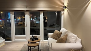 Een moderne woonkamer met een witte bank, een klein rond tafeltje en een groot raam met uitzicht op een stadsbeeld 's nachts.