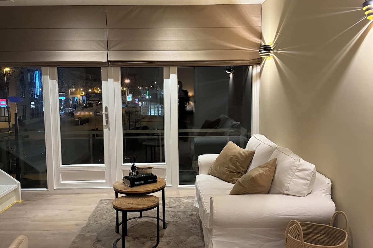 Een moderne woonkamer met een witte bank, een klein rond tafeltje en een groot raam met uitzicht op een stadsbeeld 's nachts.