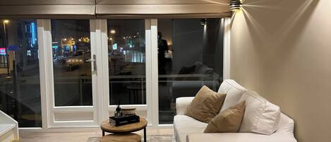 Una sala de estar moderna con un sofá blanco, una mesita redonda y un amplio ventanal con vistas a un paisaje urbano nocturno.