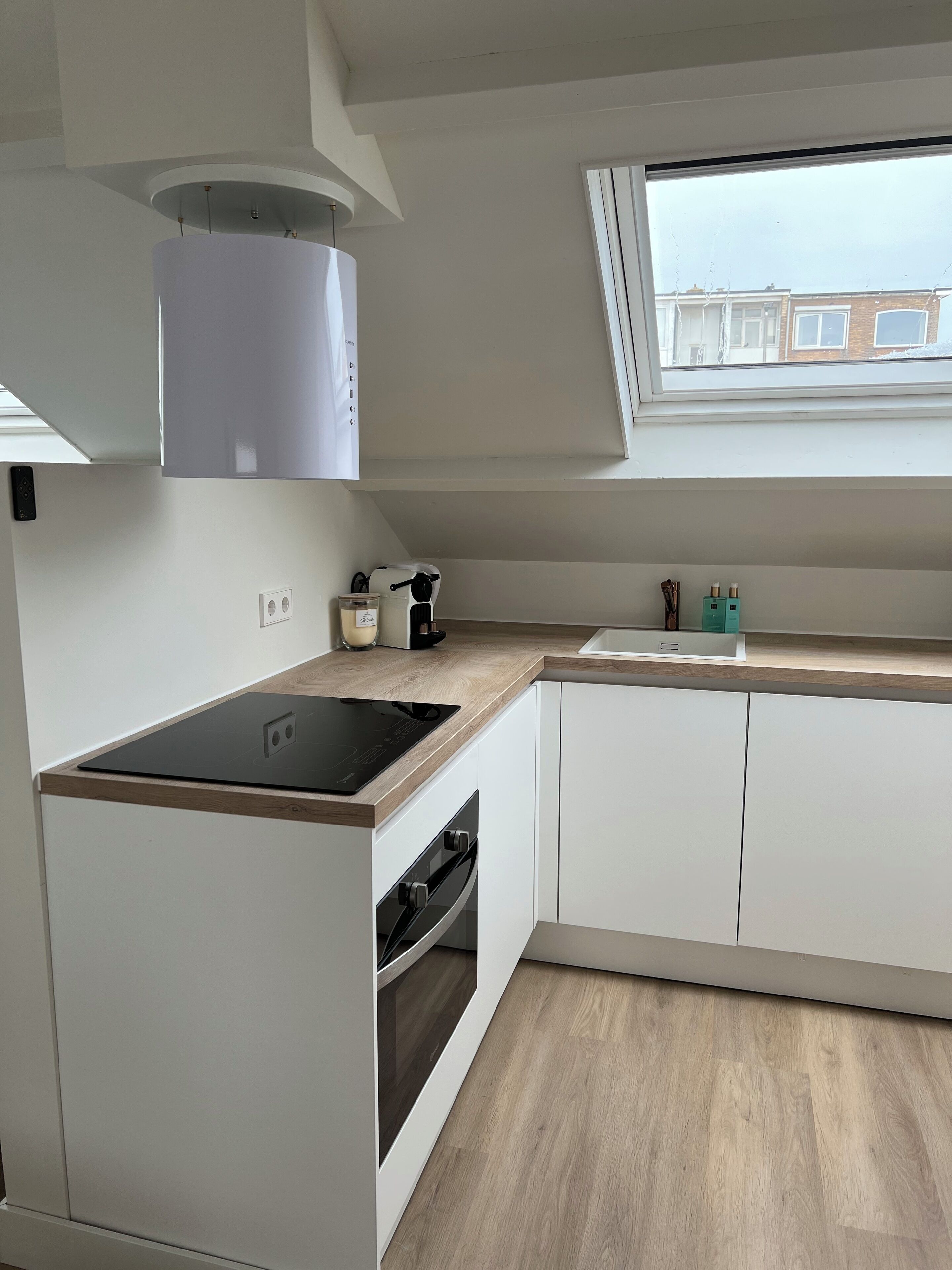 A modern kitchen with white cabinetry, a built-in oven, a black induction cooktop, a window with a view of buildings, and a light fixture above the cooktop.