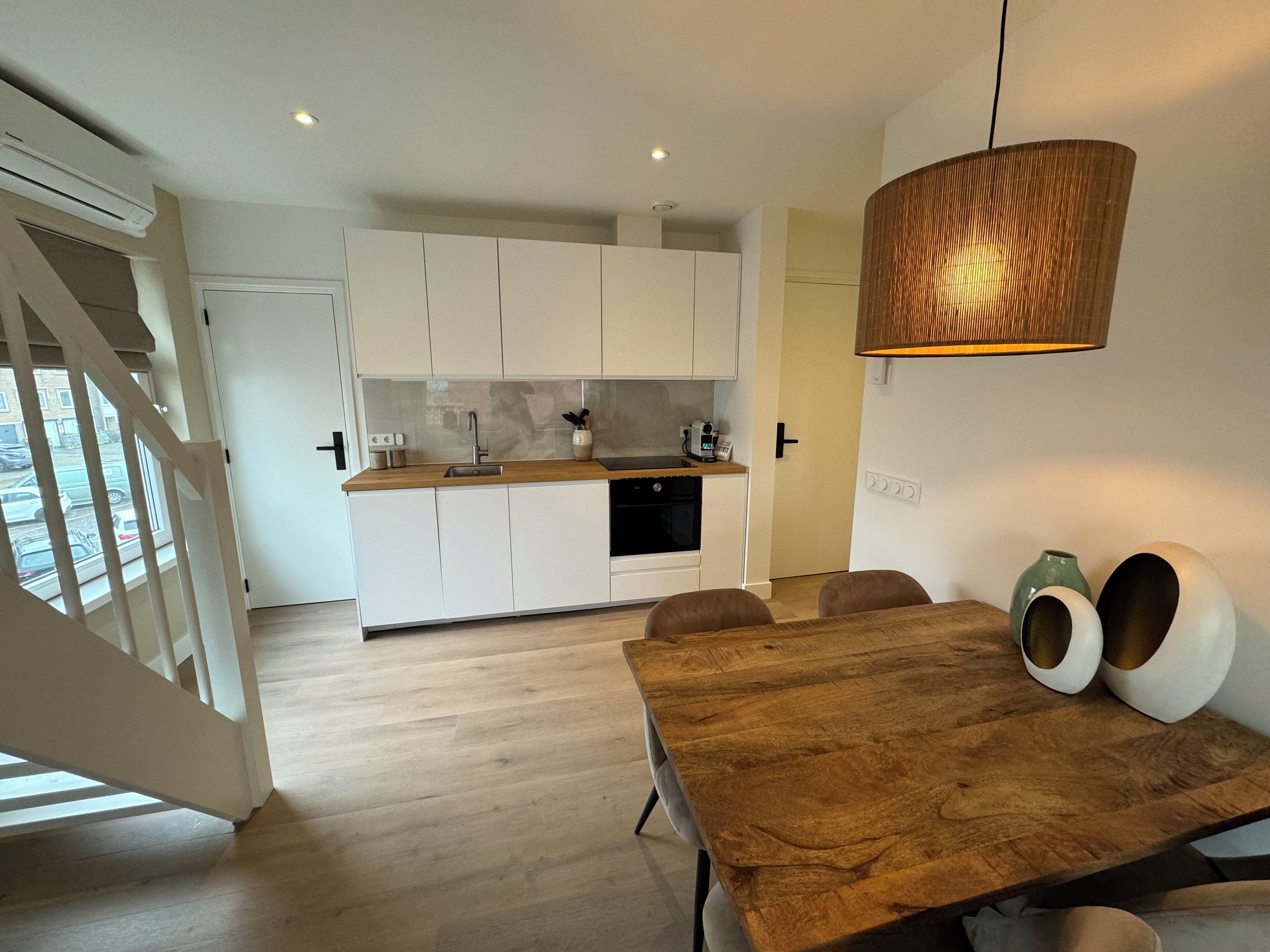 A modern kitchen with white cabinets, a wooden countertop, and a built-in oven. There is a dining area with a wooden table and chairs, and a hanging light fixture.