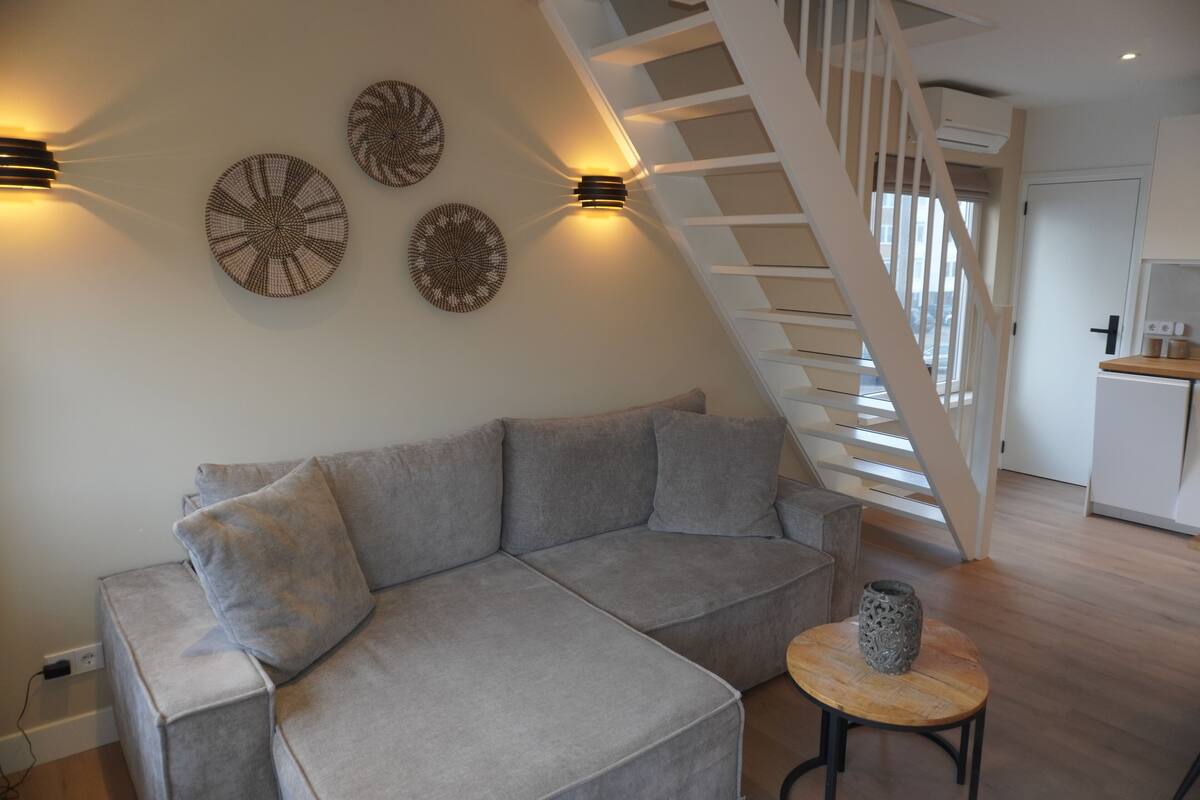 Een moderne woonkamer met een grijze modulaire bank, een klein rond tafeltje met een decoratieve vaas, en een trap die naar een hoger gelegen verdieping leidt.