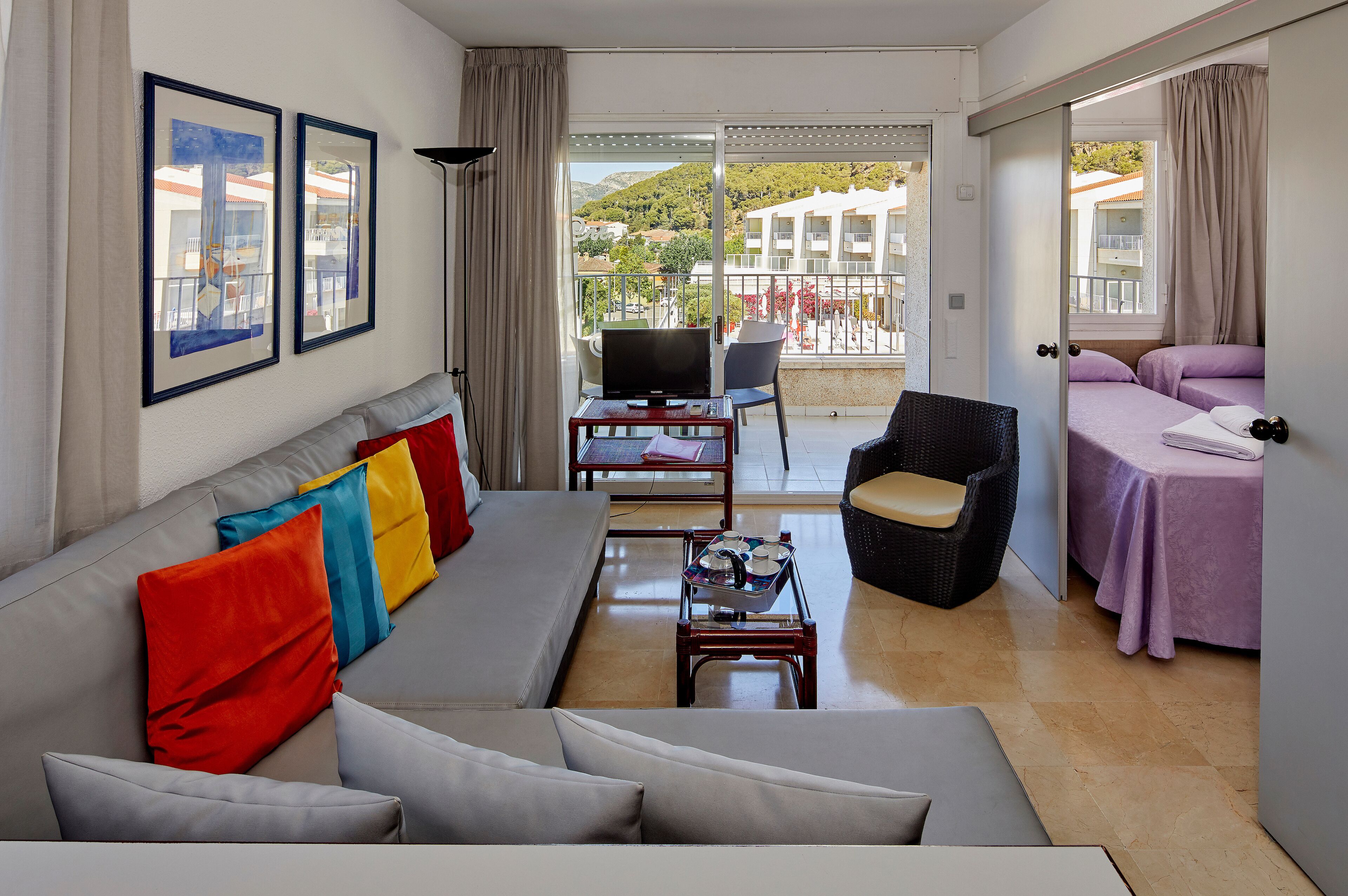 A modern living room with a sofa, colorful pillows, a TV, and a balcony with a view of buildings and hills.