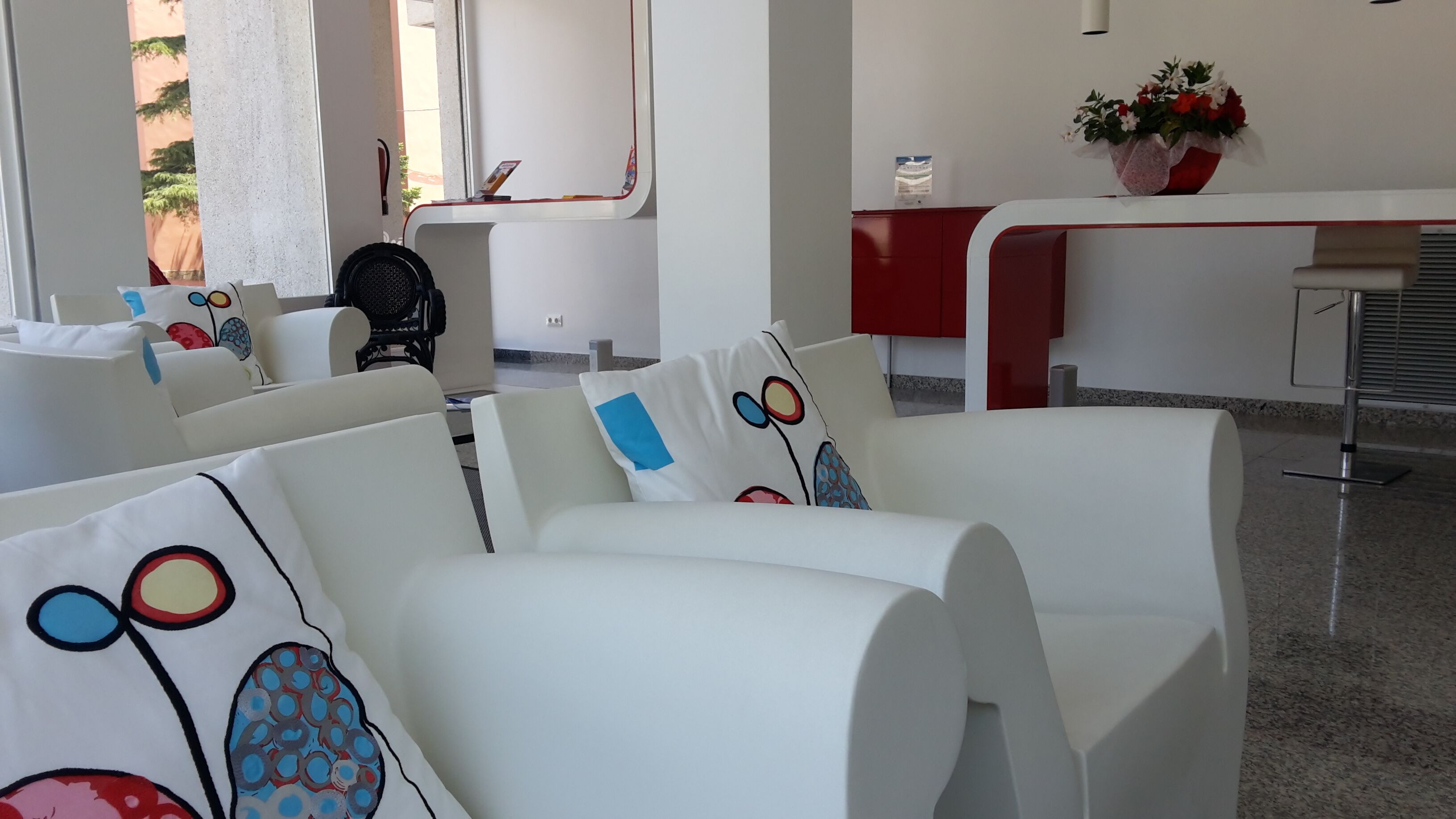 A modern hotel room with white furniture, a red reception desk, and a flower vase.