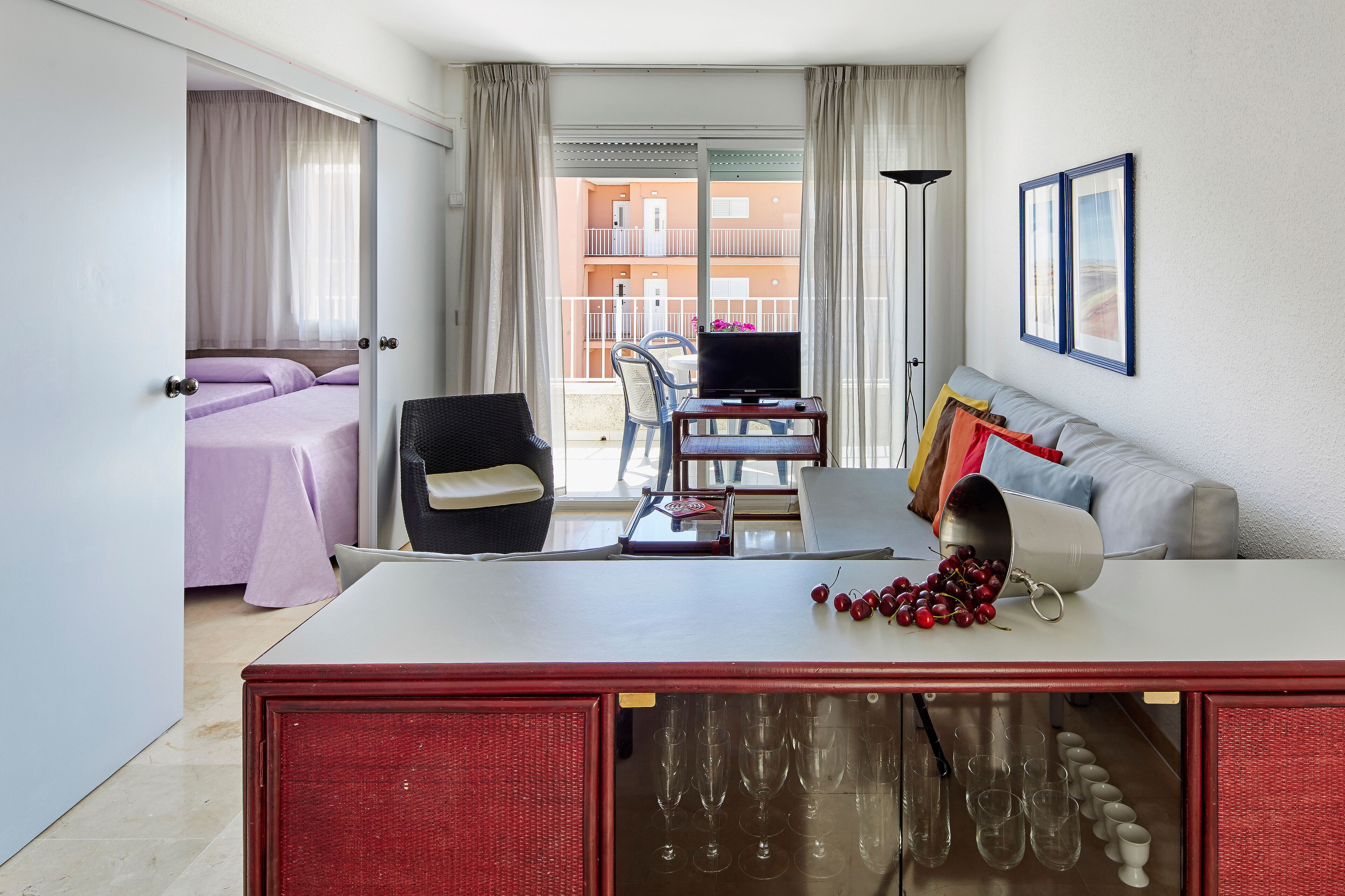 A hotel room with a bed, a table with a TV, a chair, a sofa, and a balcony with a view of buildings.