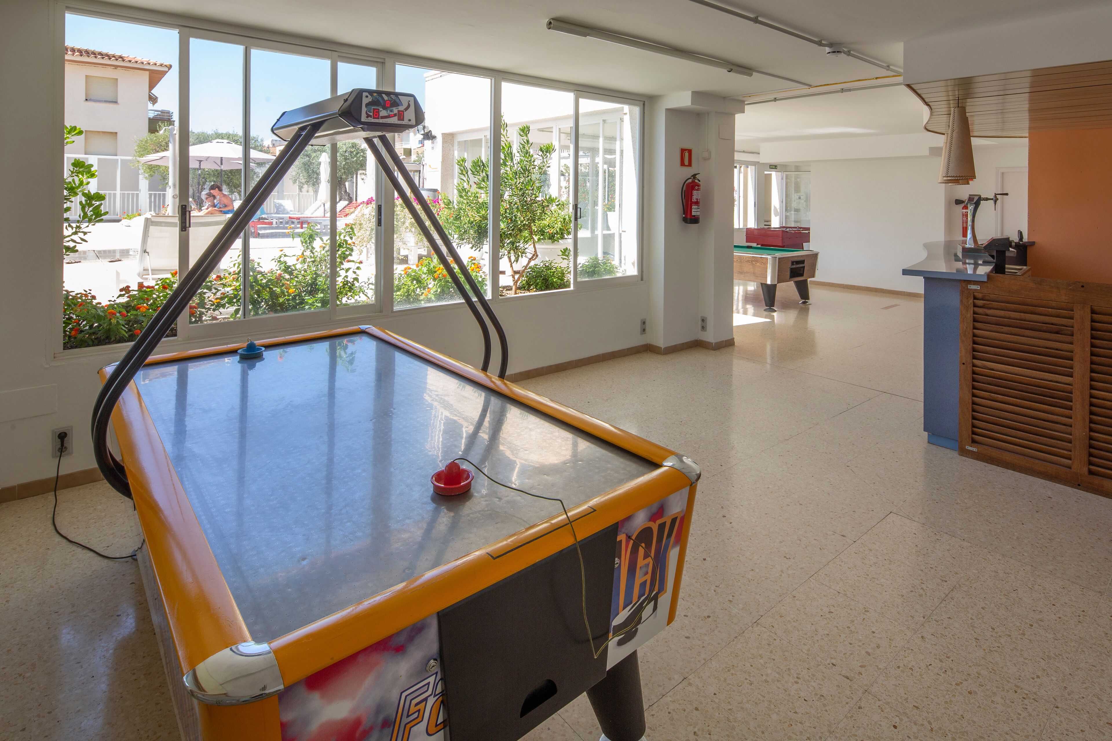 A pool table in a spacious room with large windows, a fire extinguisher on the wall, and a counter with a lamp.