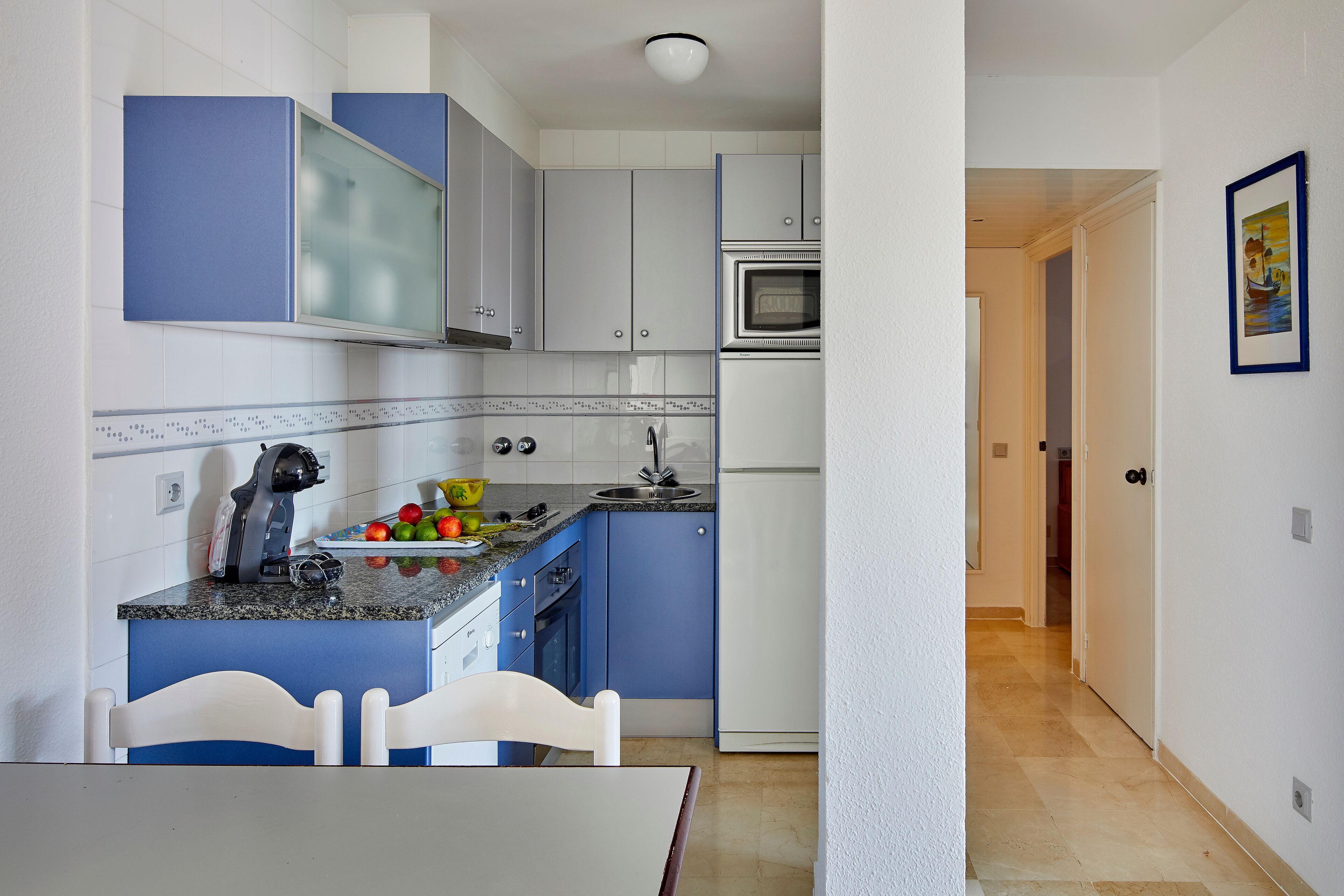 A kitchen with blue cabinets, a white refrigerator, and a microwave. There is a dining table with chairs and a fruit bowl on the counter.