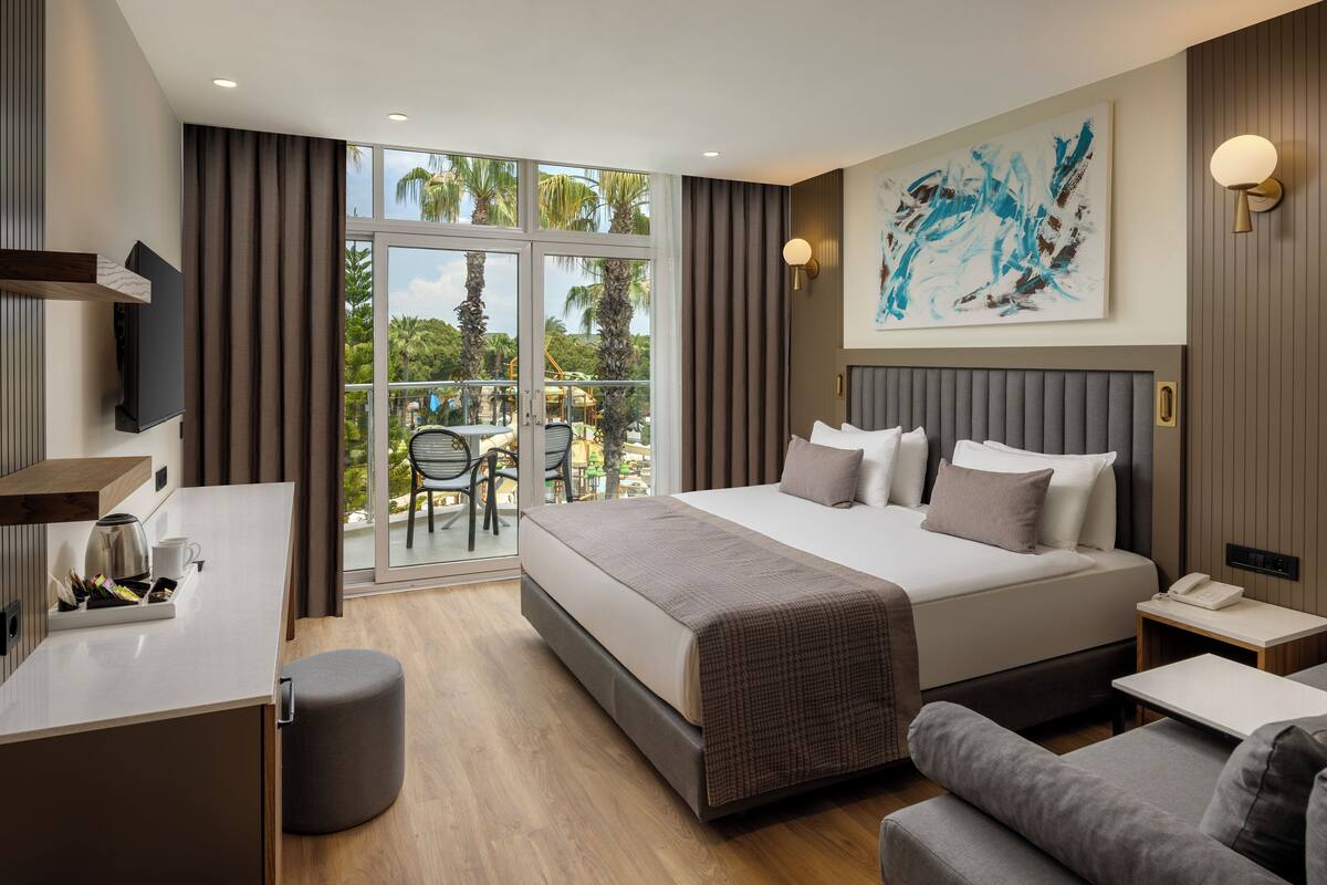 A modern hotel room with a large bed, a desk, a sofa, and a balcony with a view of palm trees.