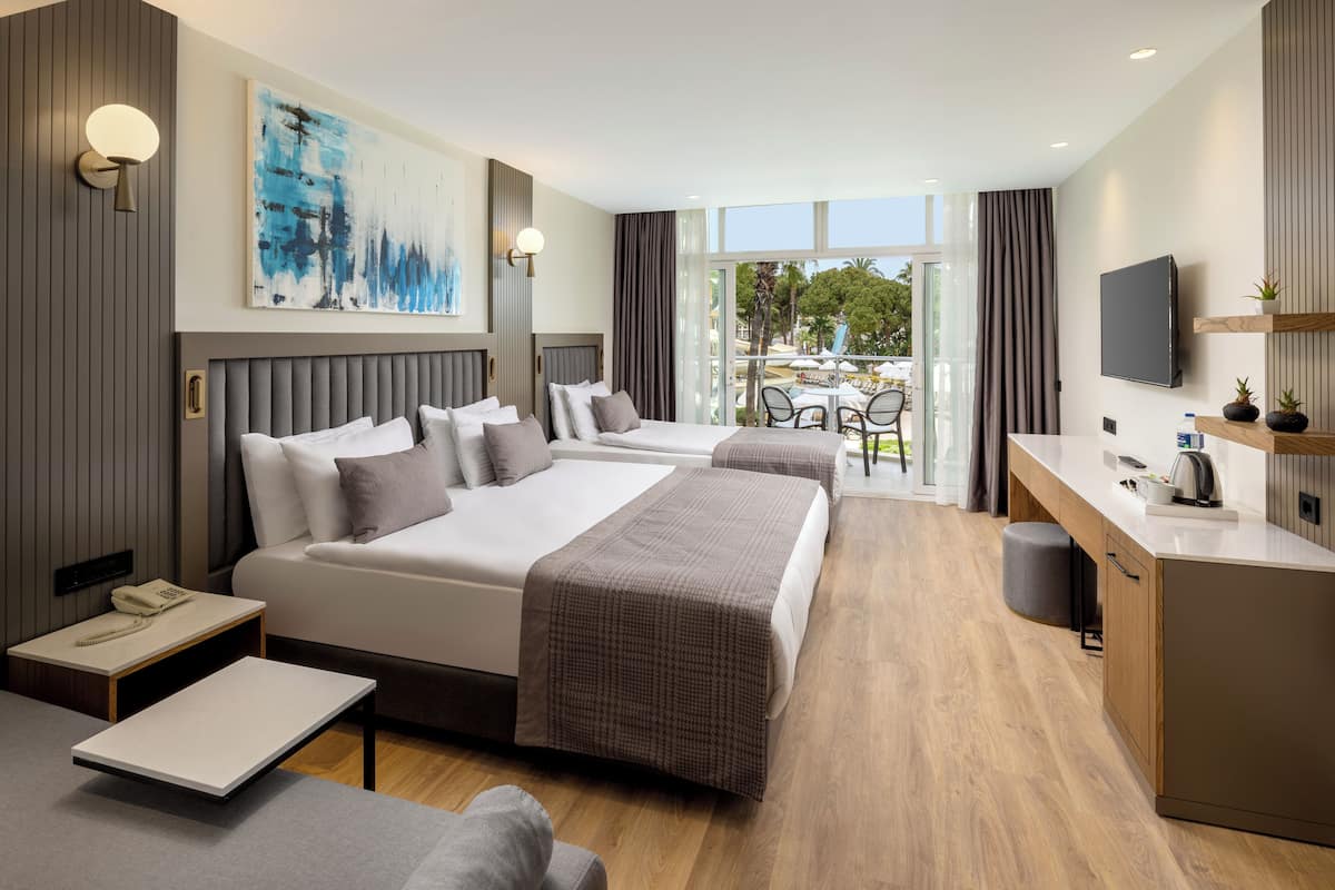 A modern hotel room with two beds, a desk, a flat-screen TV, and a balcony with outdoor seating.