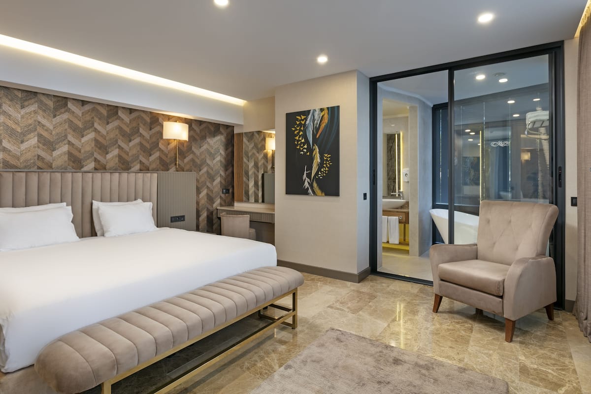 A modern hotel room with a large bed, a bench, a chair, and a glass door leading to a bathroom.