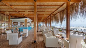 A covered outdoor seating area with wooden flooring and white chairs, overlooking a beach with umbrellas and a bar area in the background. - DOBEDAN WORLD PALACE - All inclusive (Kemer)