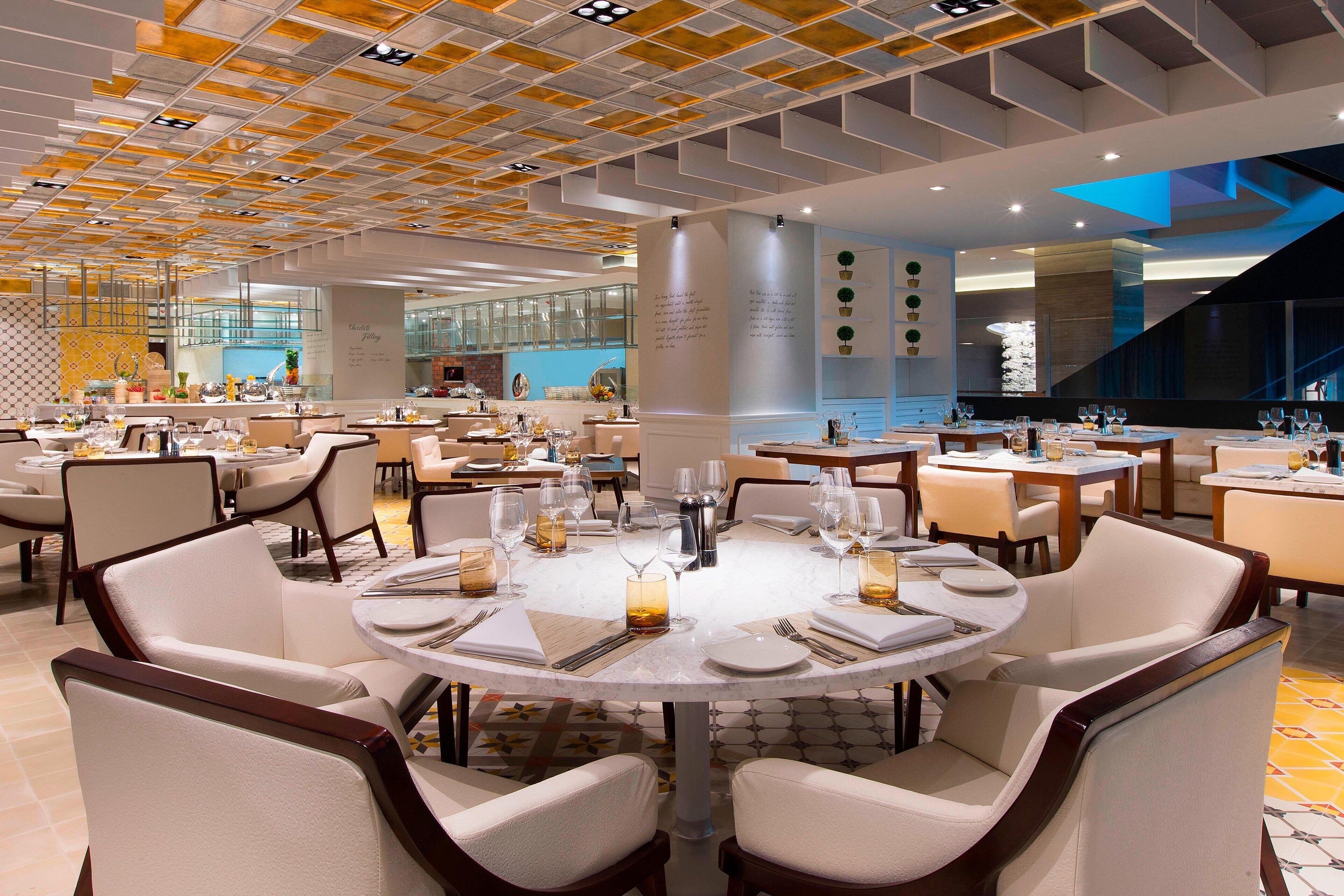 A spacious dining area with round tables and white chairs, featuring a modern ceiling design and a visible kitchen area in the background.