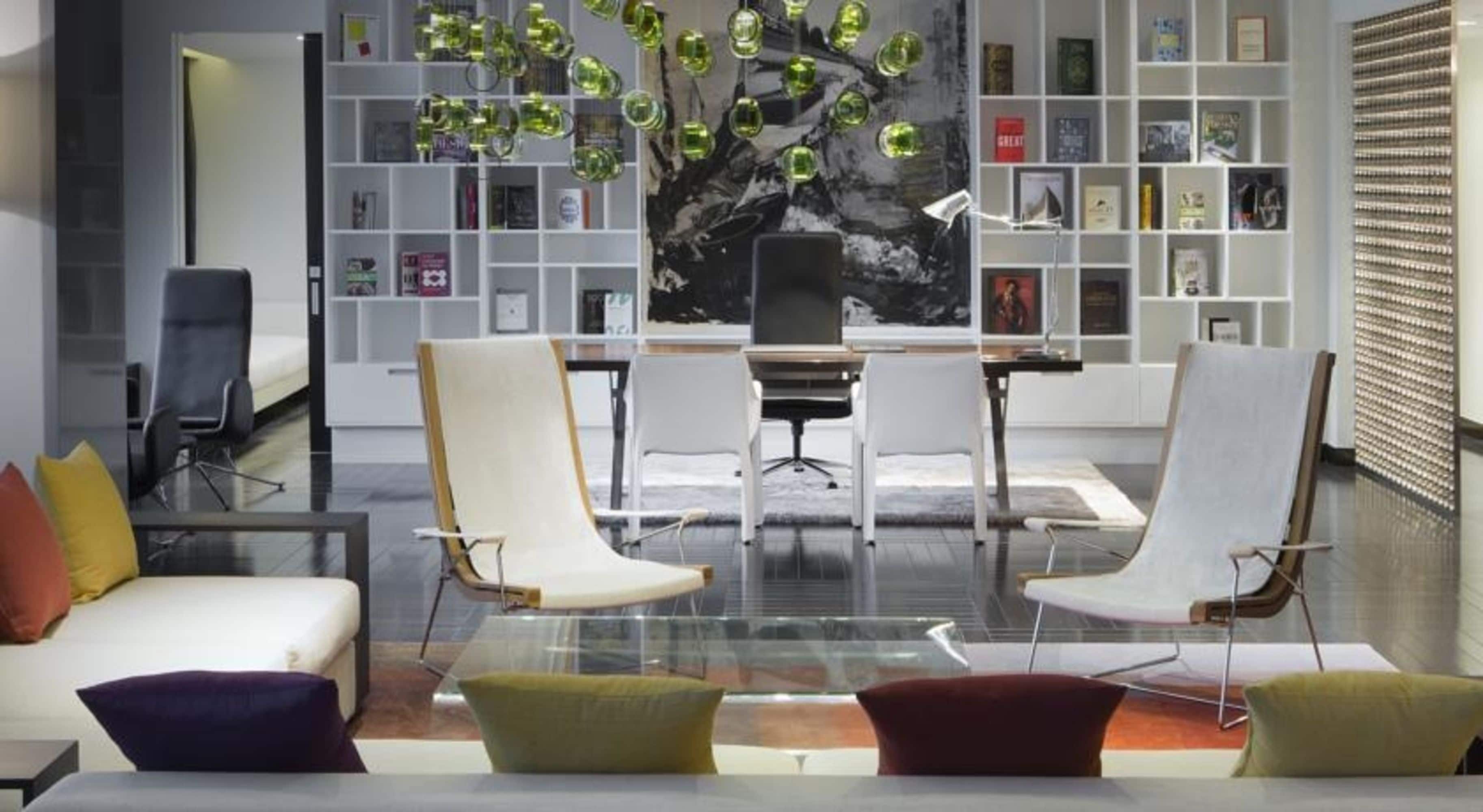 A modern library with a glass table, two white armchairs, and a bookshelf filled with books.