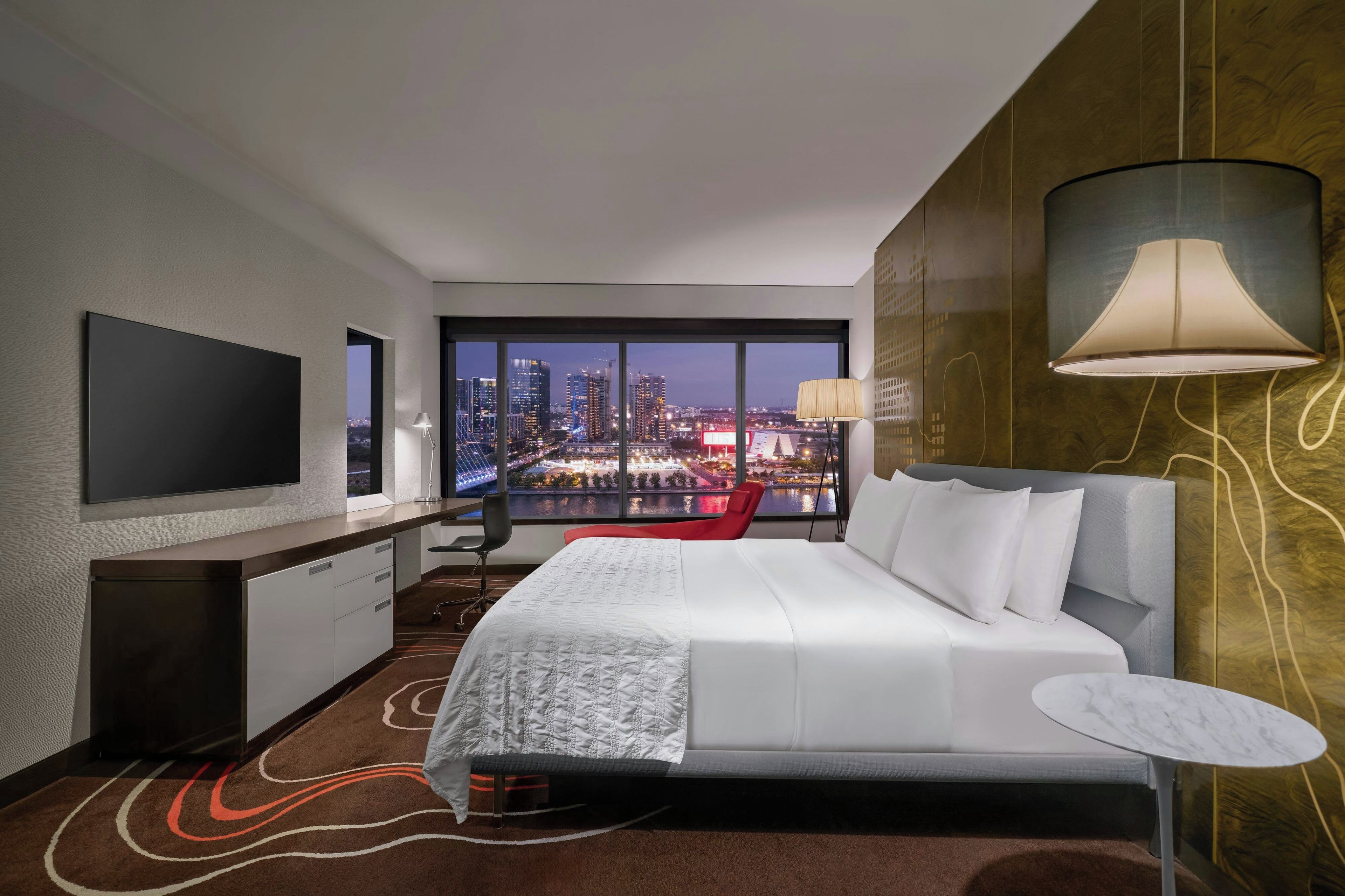 A hotel room with a large bed, a TV, a desk, and a view of the cityscape.