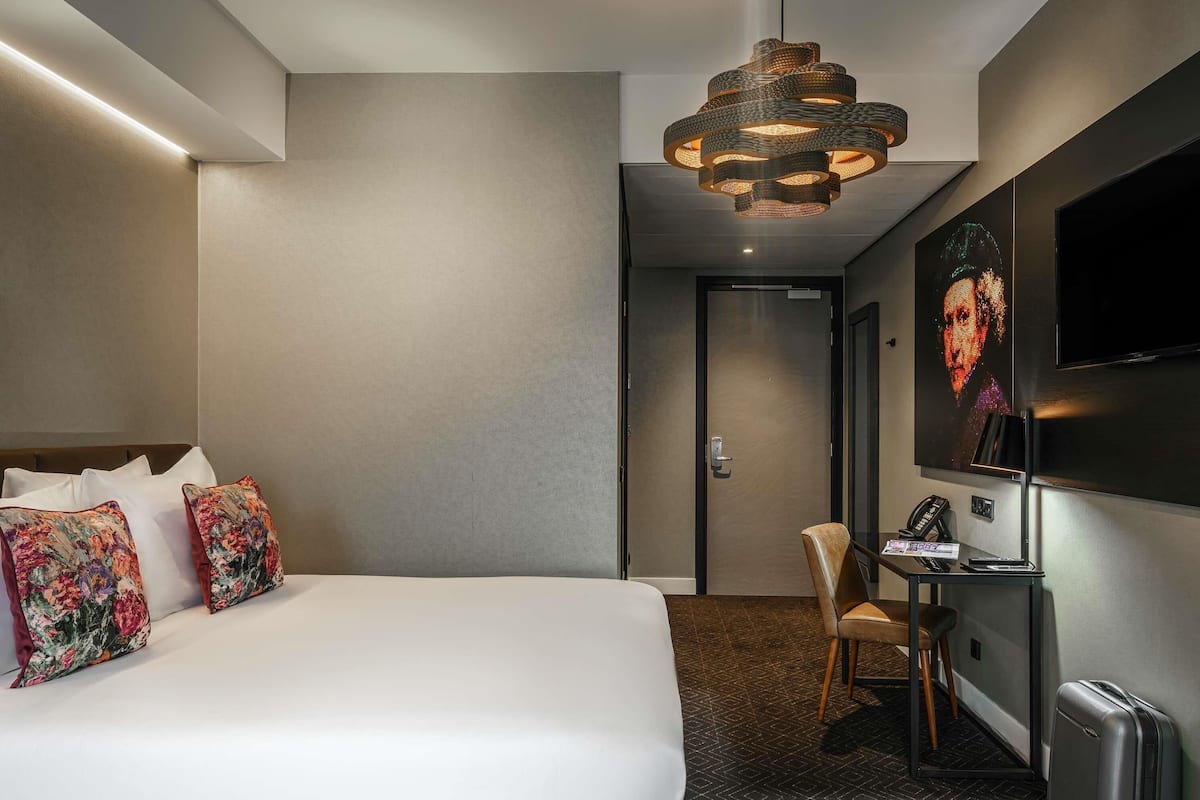 A hotel room with a large bed, a desk with a chair, a television, and a modern chandelier.