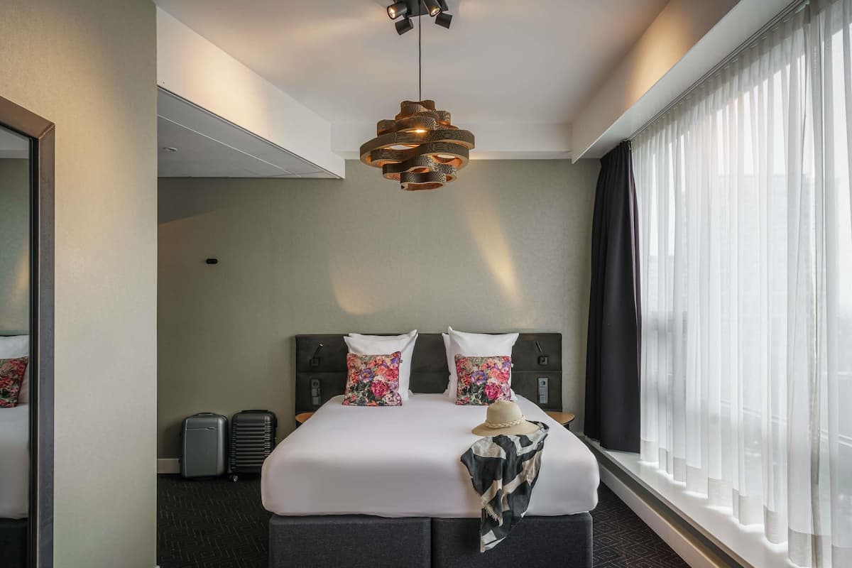 A hotel room with a large bed, a modern chandelier, a large window with sheer curtains, and a small table with a lamp.