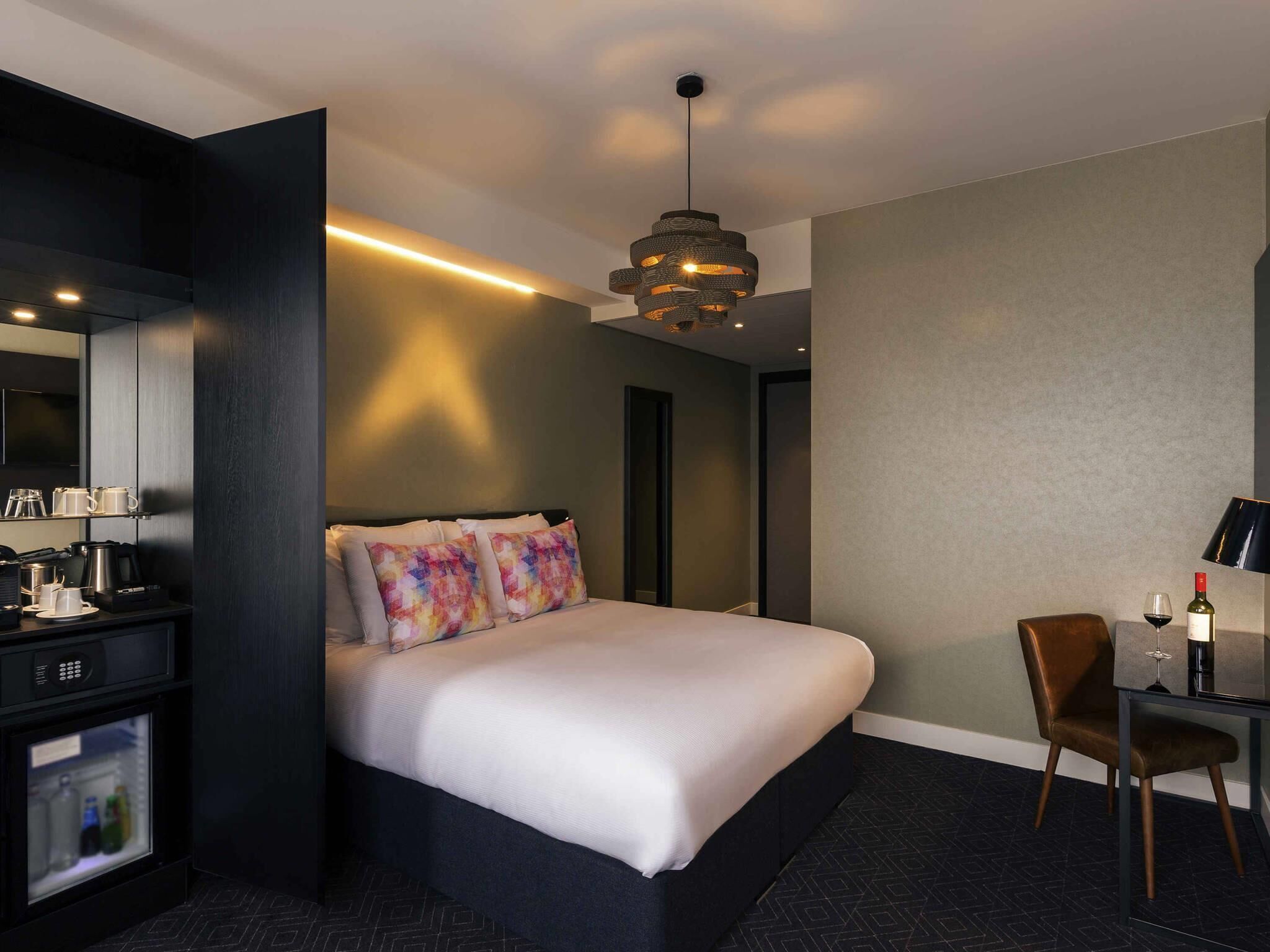 A hotel room with a bed, a desk, a chair, a minibar, and a hanging light fixture.