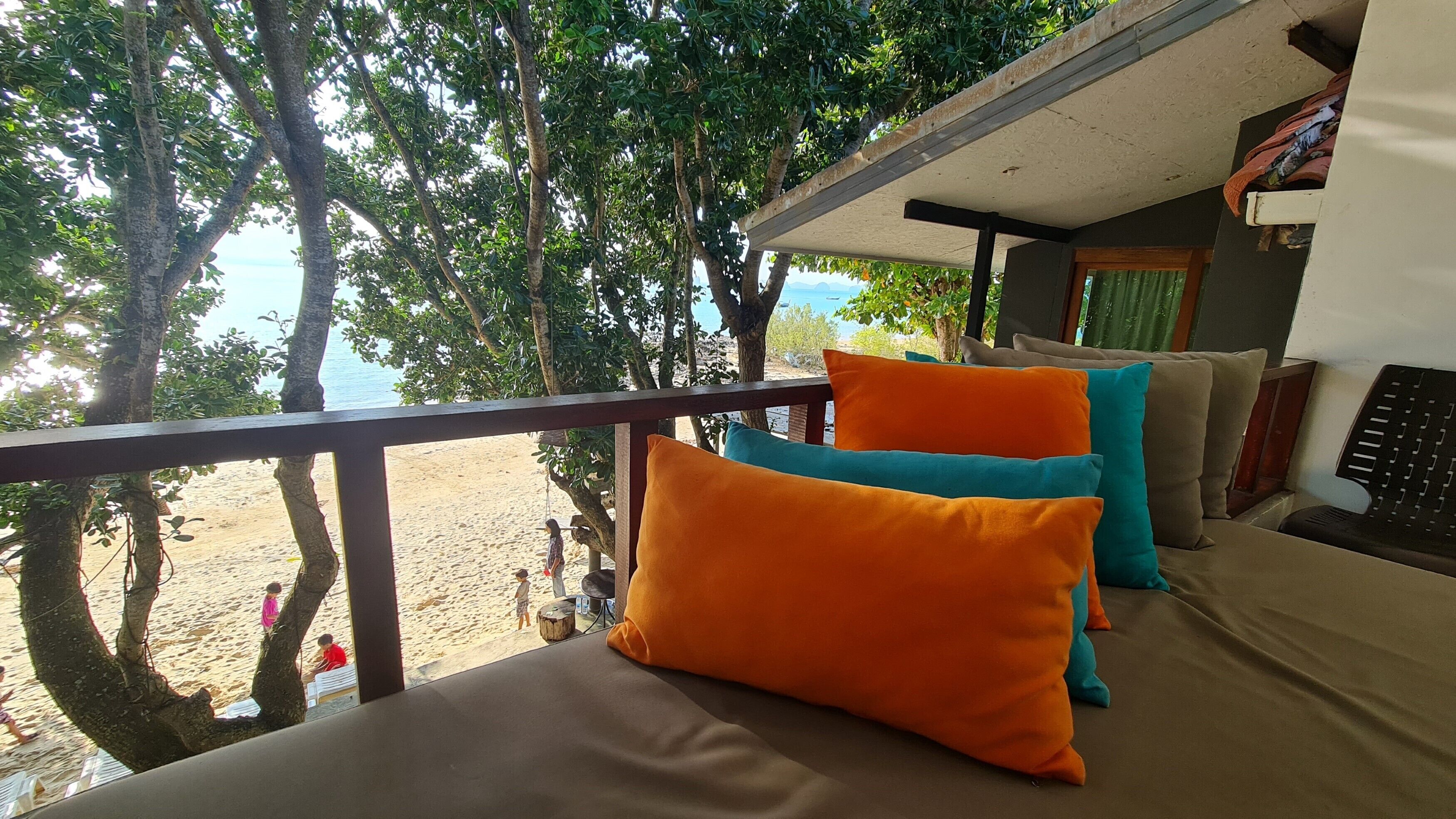 A balcony with a view of the beach, featuring a sofa with orange cushions and a wooden railing.