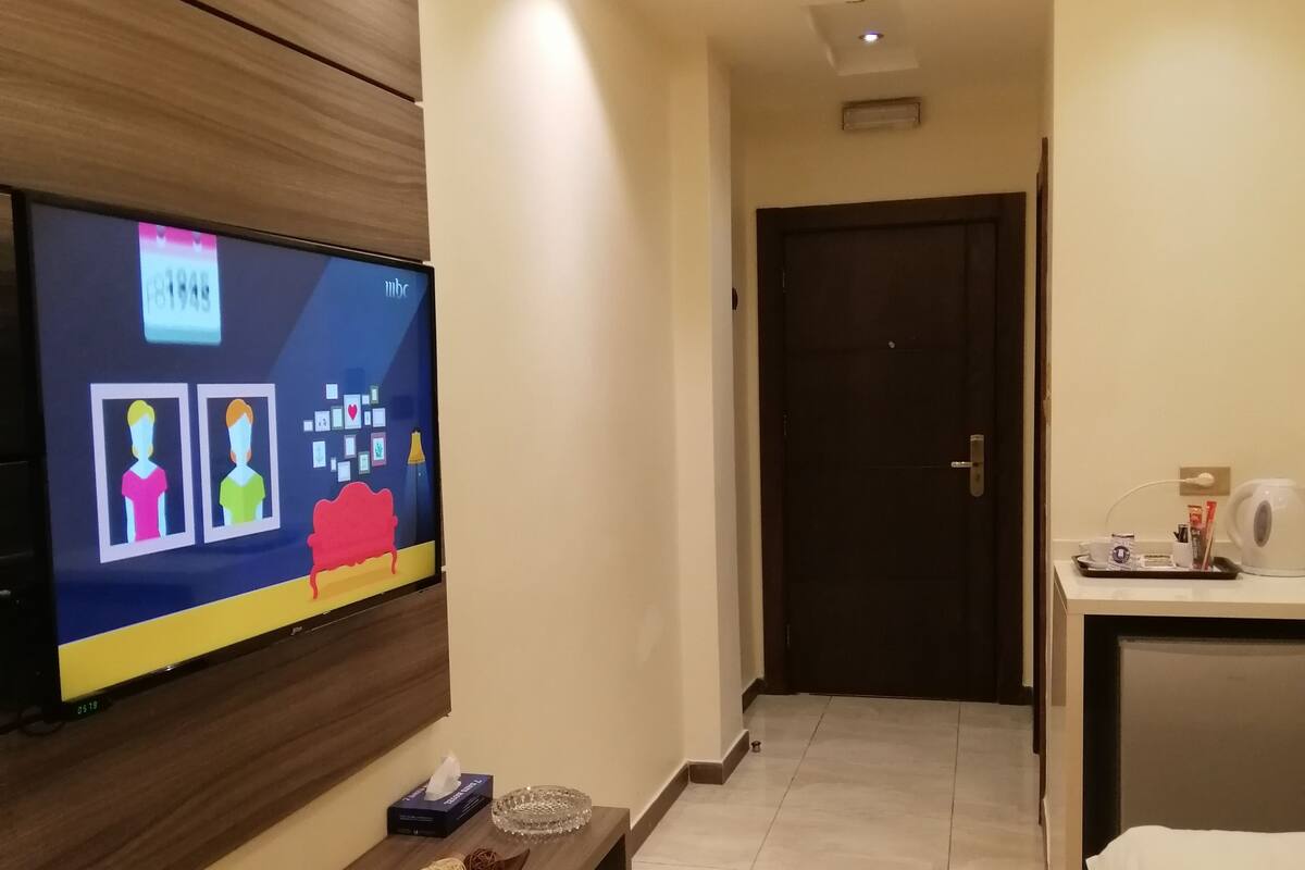 Habitació d'hotel amb televisió de pantalla plana penjada a la paret, taula consola de fusta, llit amb roba de llit blanca, escriptori amb una tetera i una porta al fons.