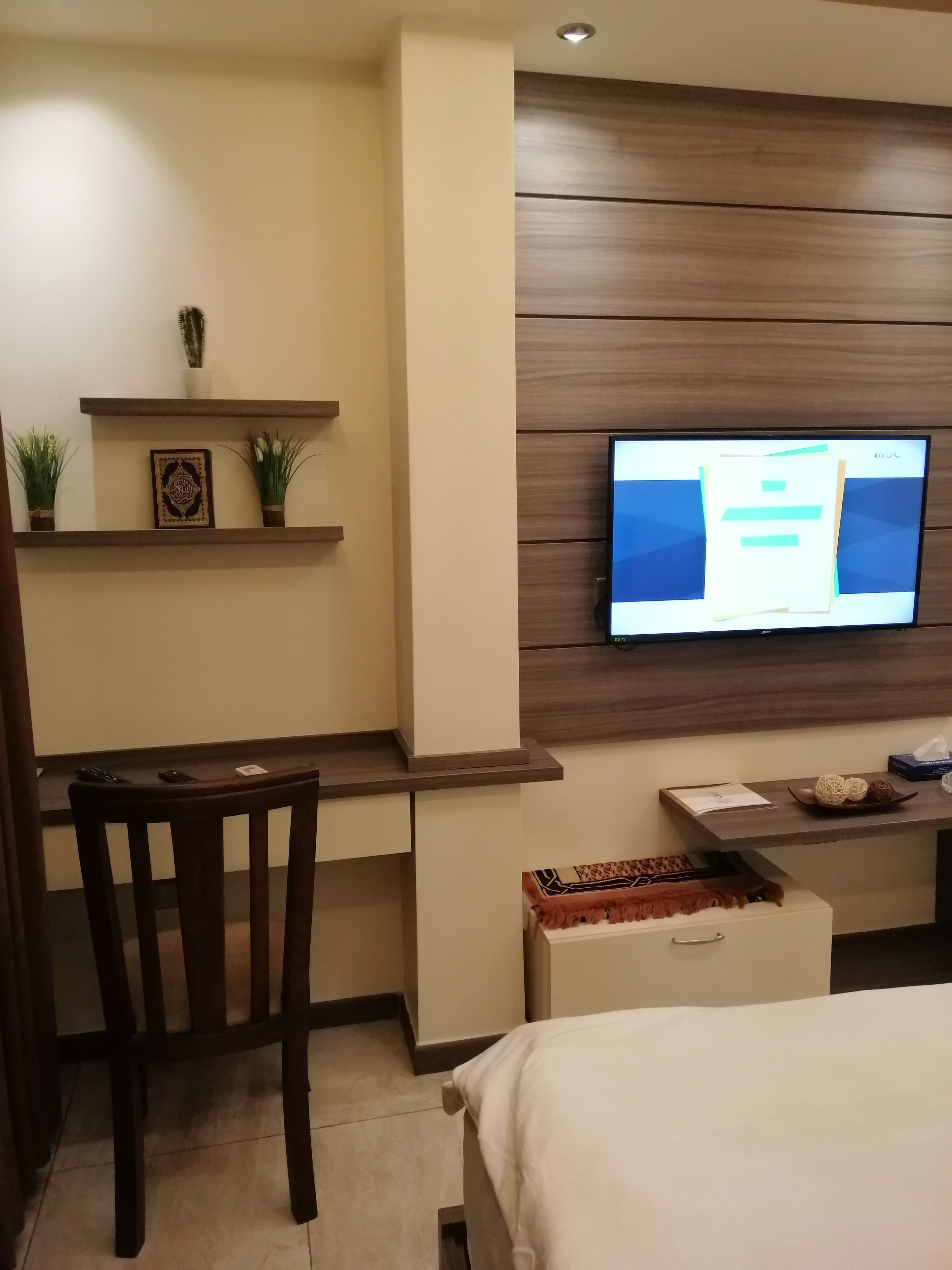 A modern hotel room with a wooden desk, a television mounted on the wall, a bed, and a small shelf with decorative items.