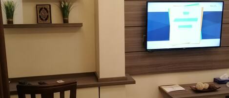 A modern hotel room with a wooden desk, a television mounted on the wall, a bed, and a small shelf with decorative items.