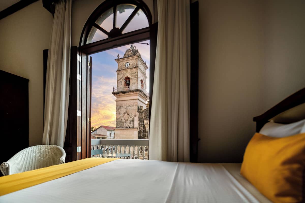 Quarto de hotel com uma cama, vista para uma torre histórica e varanda.